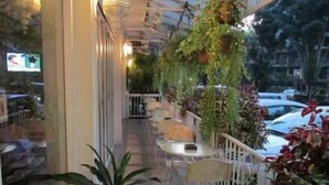 Un café al aire libre con mesas y sillas, rodeado de plantas en macetas y vistas a coches estacionados.