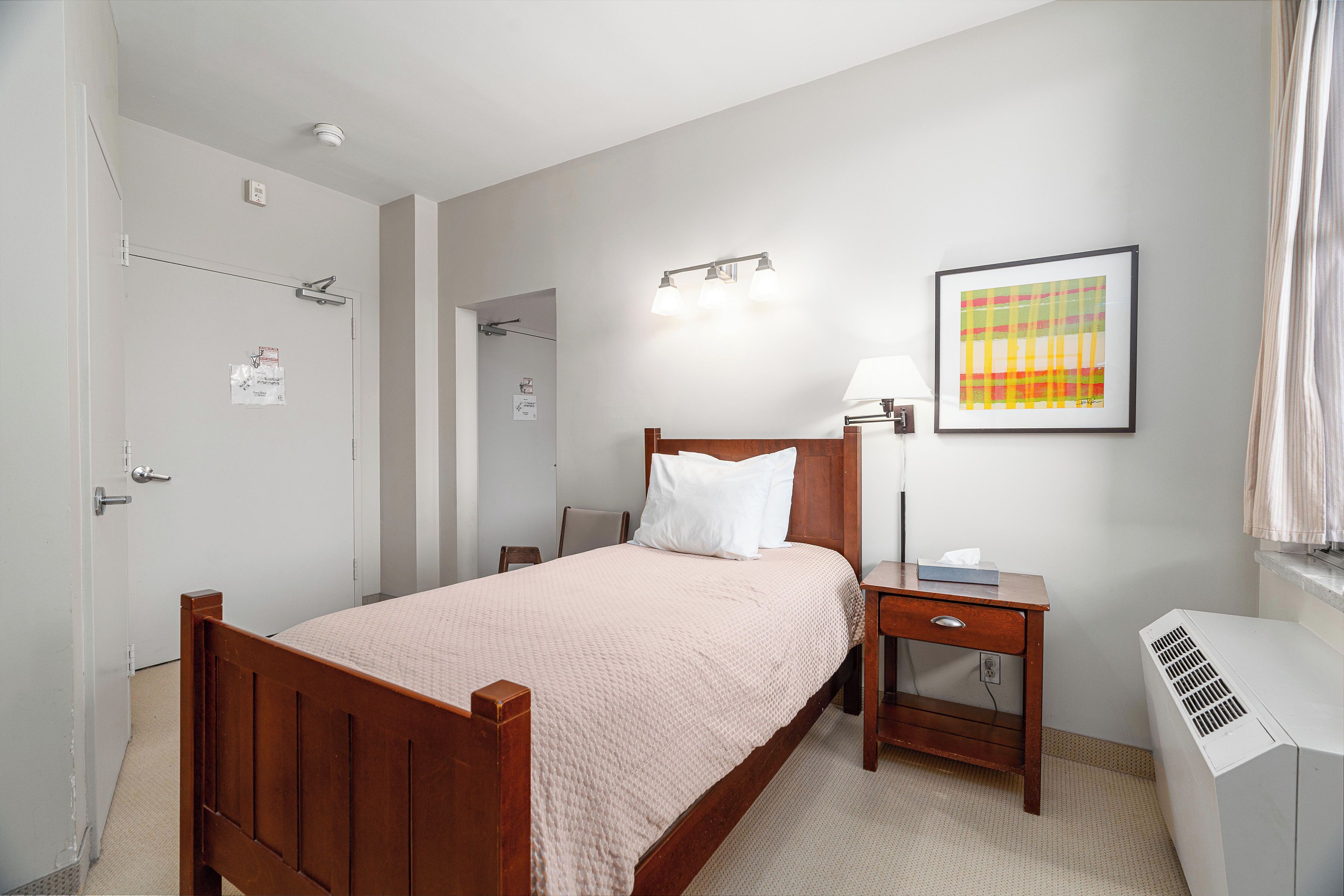 Une chambre à coucher avec un lit en bois, une table de chevet, un bureau, une lampe fixée au mur et un radiateur.