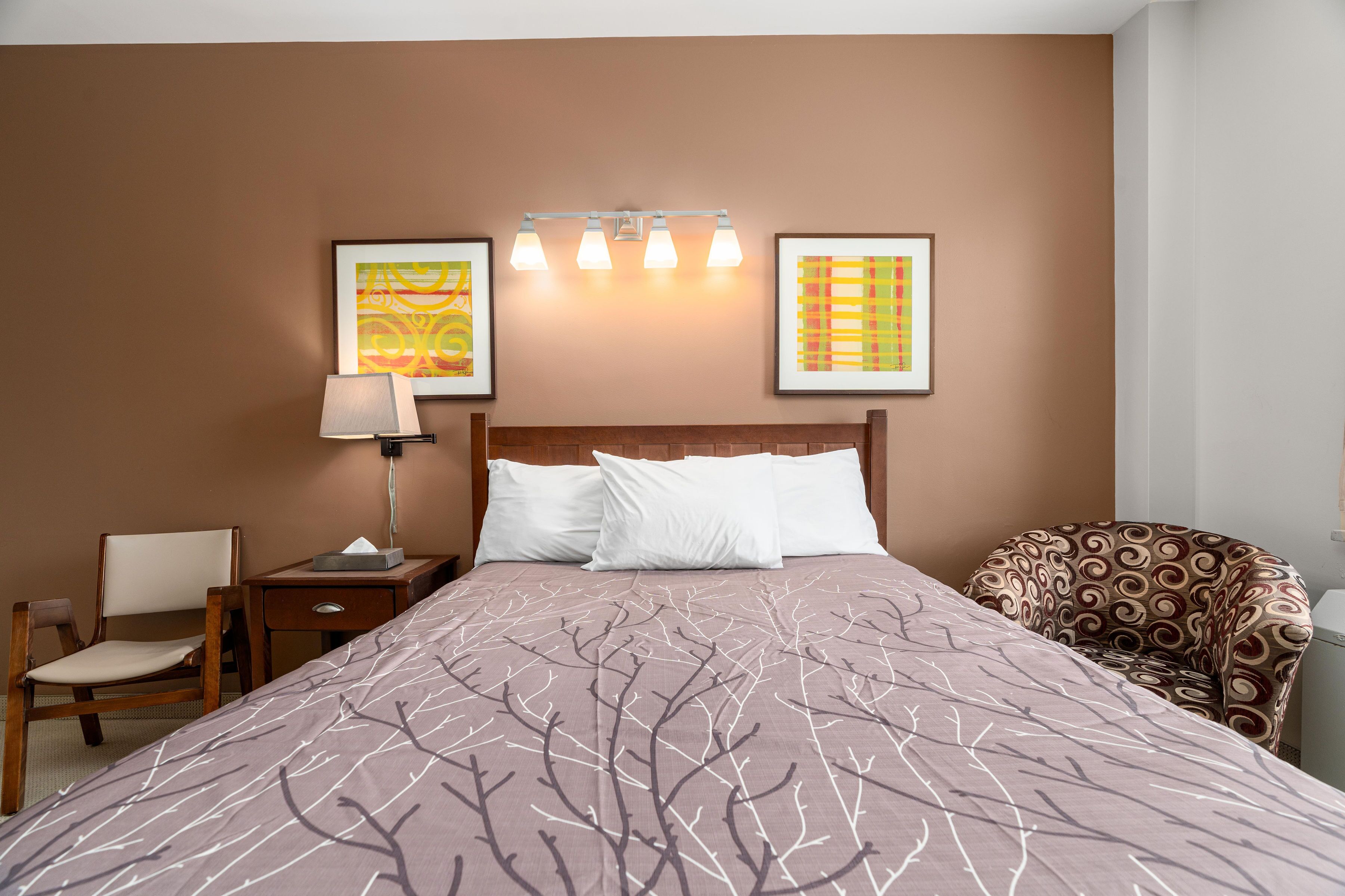 A hotel room with a large bed, a nightstand, a chair, and two wall-mounted light fixtures.