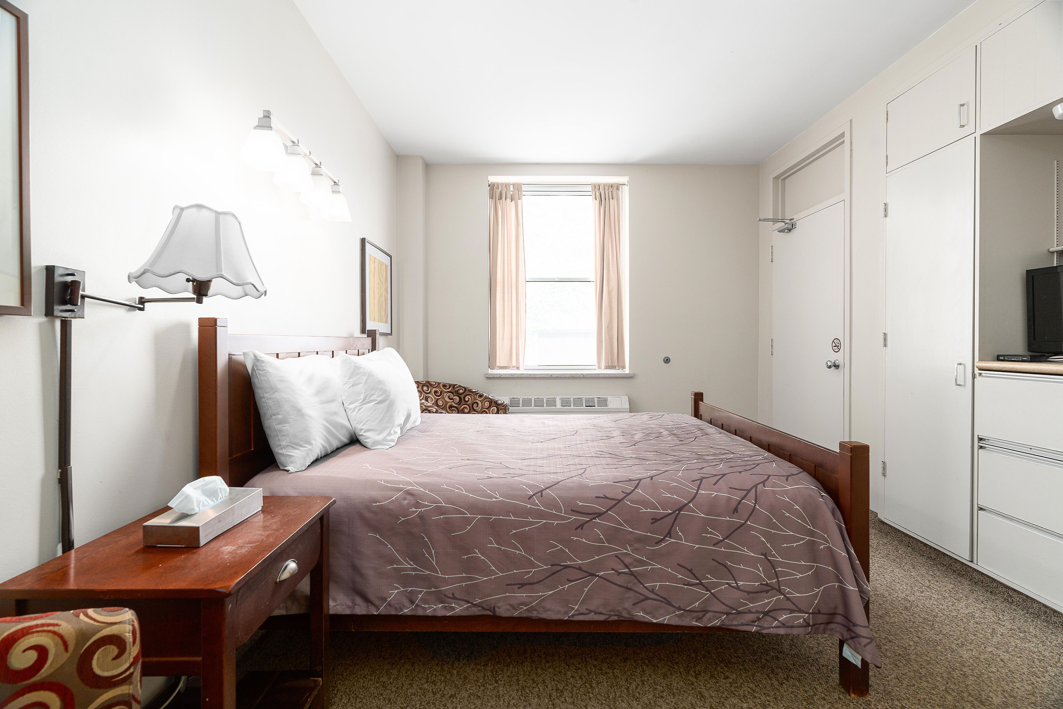 Une chambre à coucher comprenant un lit, une table de chevet, une lampe, une fenêtre et une armoire.