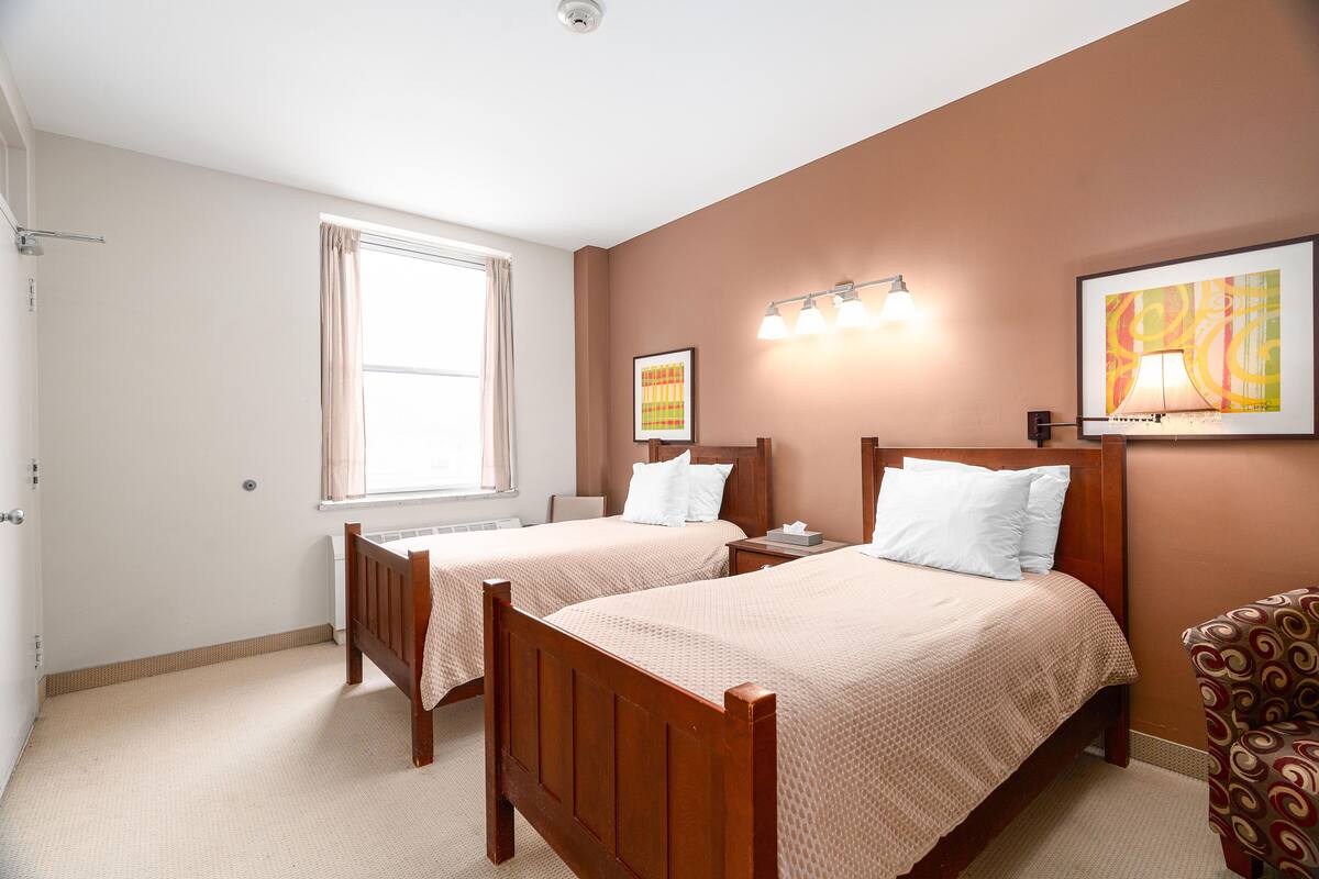 Une chambre d’hôtel avec deux lits en bois, une fenêtre avec des rideaux, un luminaire fixé au mur et un tableau encadré.