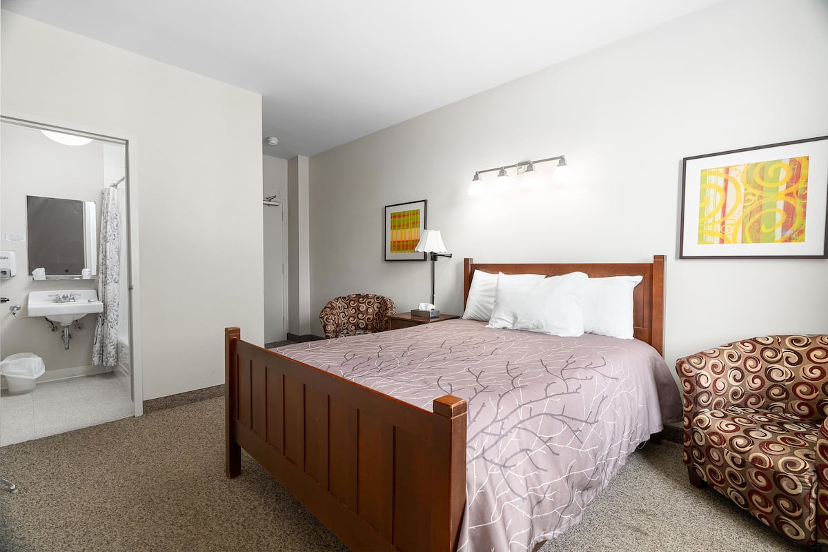Une chambre à coucher avec un lit en bois, une chaise, une table de chevet, une lampe et une salle de bain visible à travers une porte ouverte.