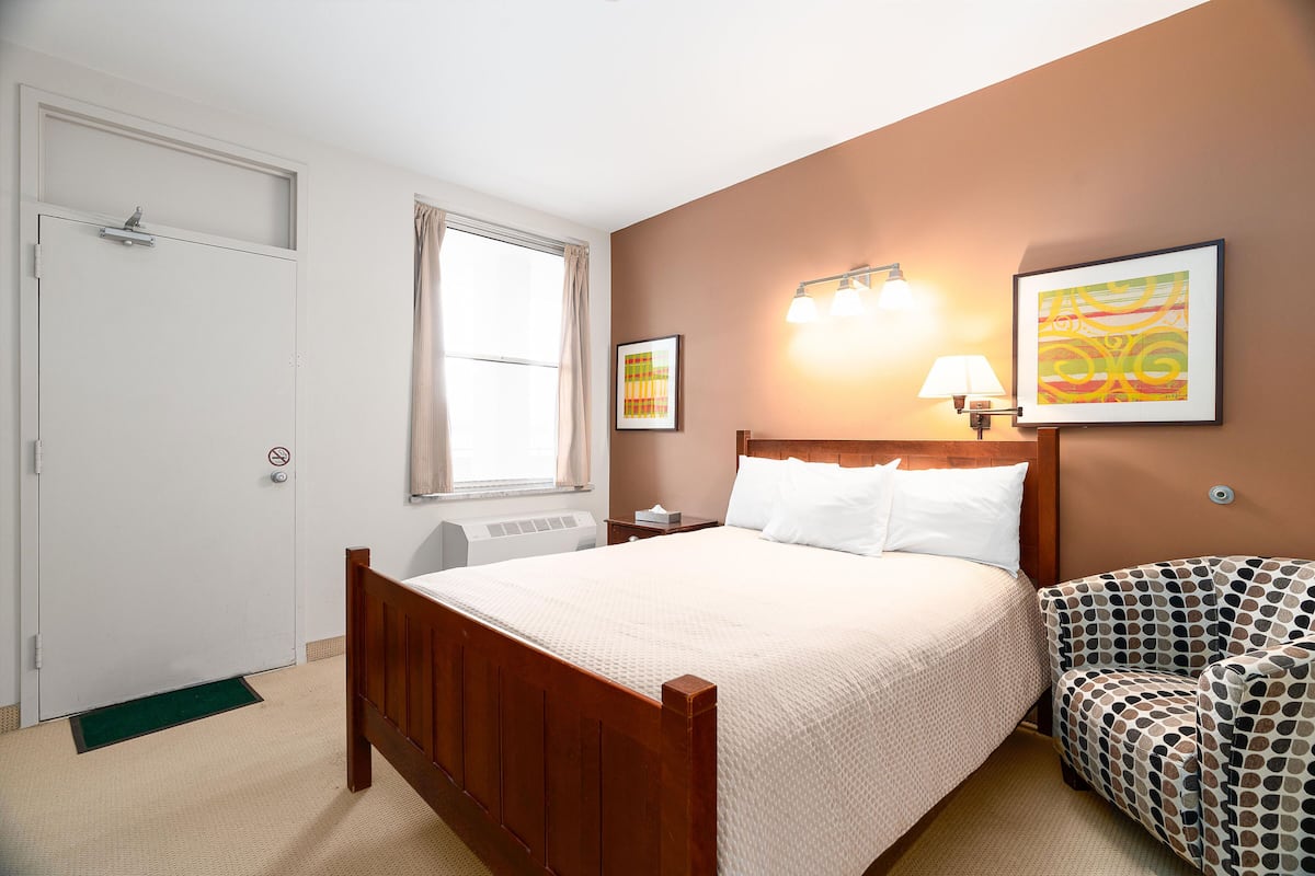 Une chambre d’hôtel avec un lit en bois, une table de chevet, une lampe, une chaise et des œuvres d’art accrochées aux murs.