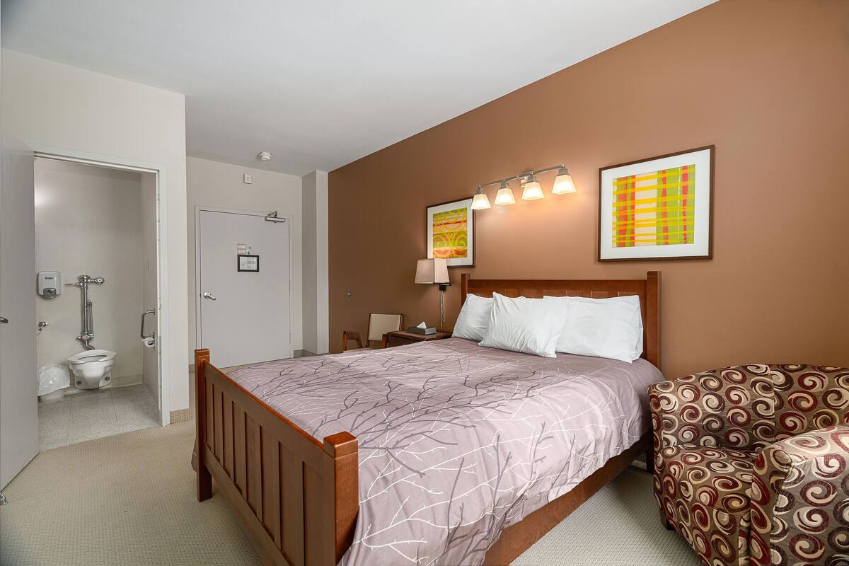 Une chambre à coucher avec un lit en bois, un fauteuil à motifs, une table de chevet et deux lampes fixées au mur.