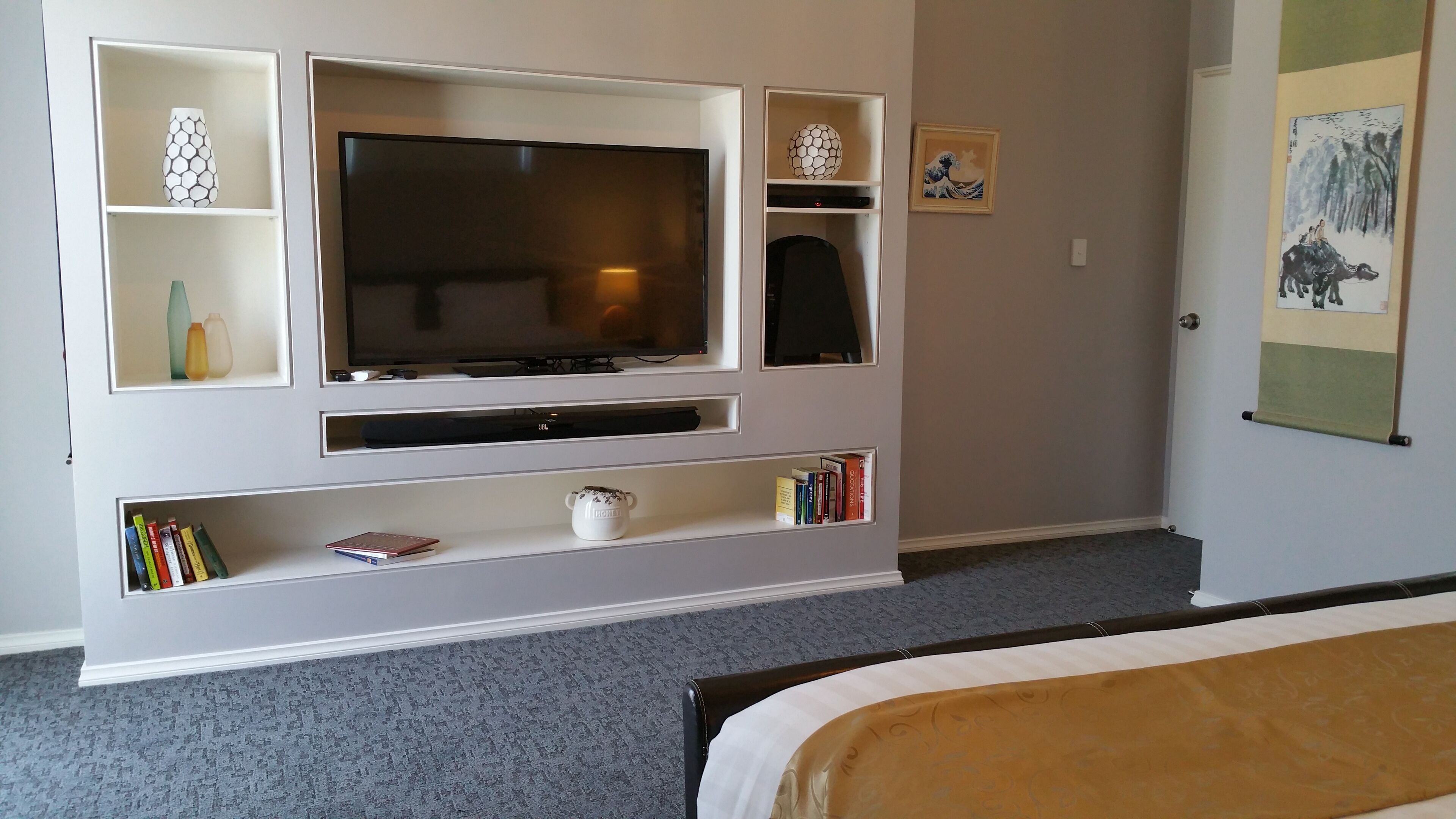 A hotel room with a flat-screen TV mounted on a white wall, a bookshelf with books and decorative items, a bed with a white and brown bedspread, and a framed artwork on the wall.