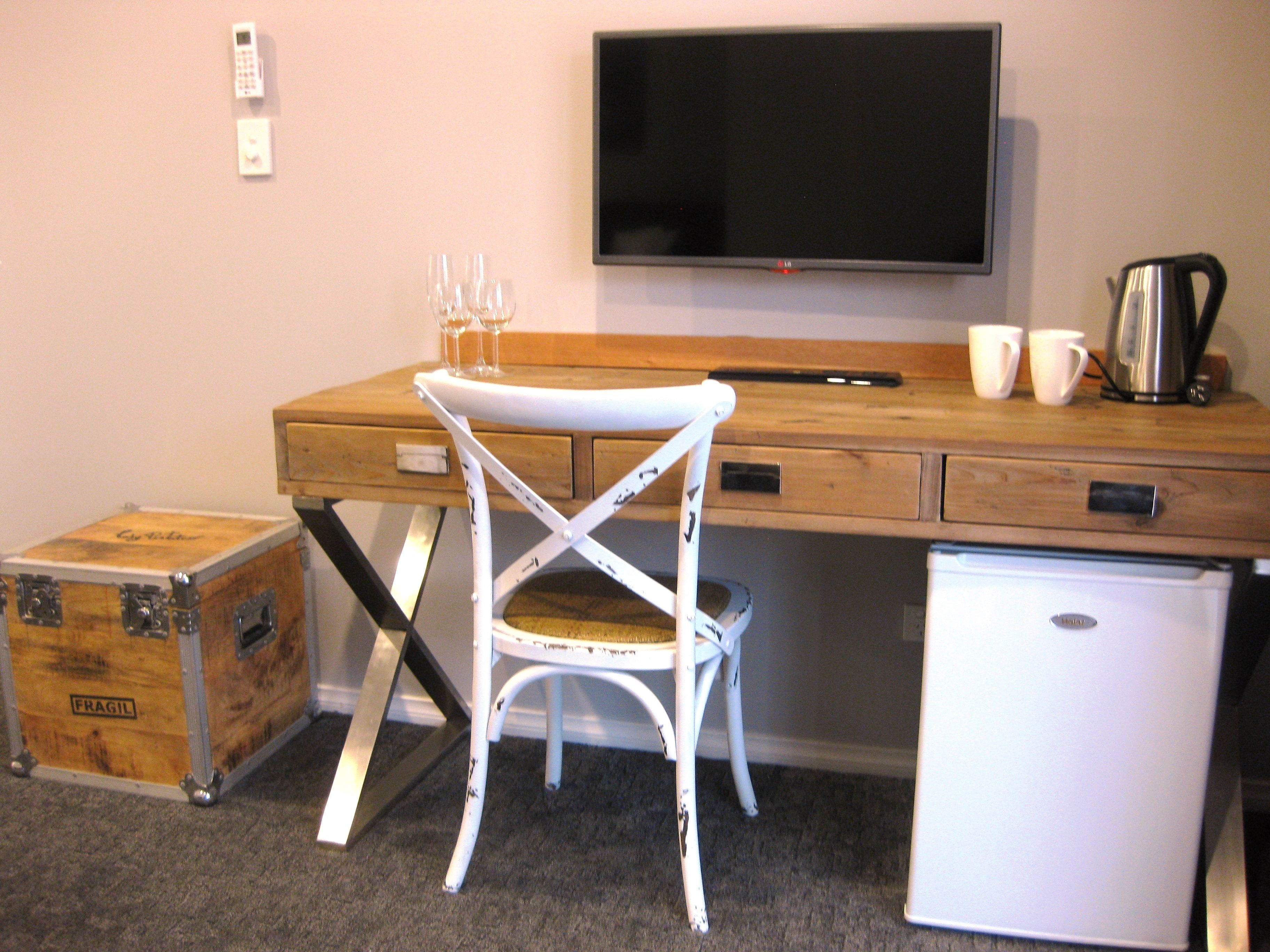 Una habitación compacta con un escritorio de madera, una silla blanca, un refrigerador pequeño y un televisor en la pared.