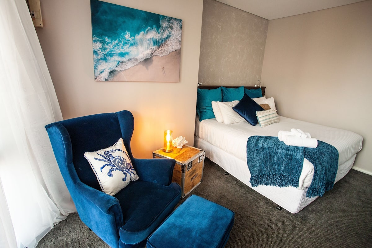 Ein Schlafzimmer mit einem Bett, einem blauen Sessel, einer kleinen Holzschatulle und einem Gemälde mit einer Strandansicht.