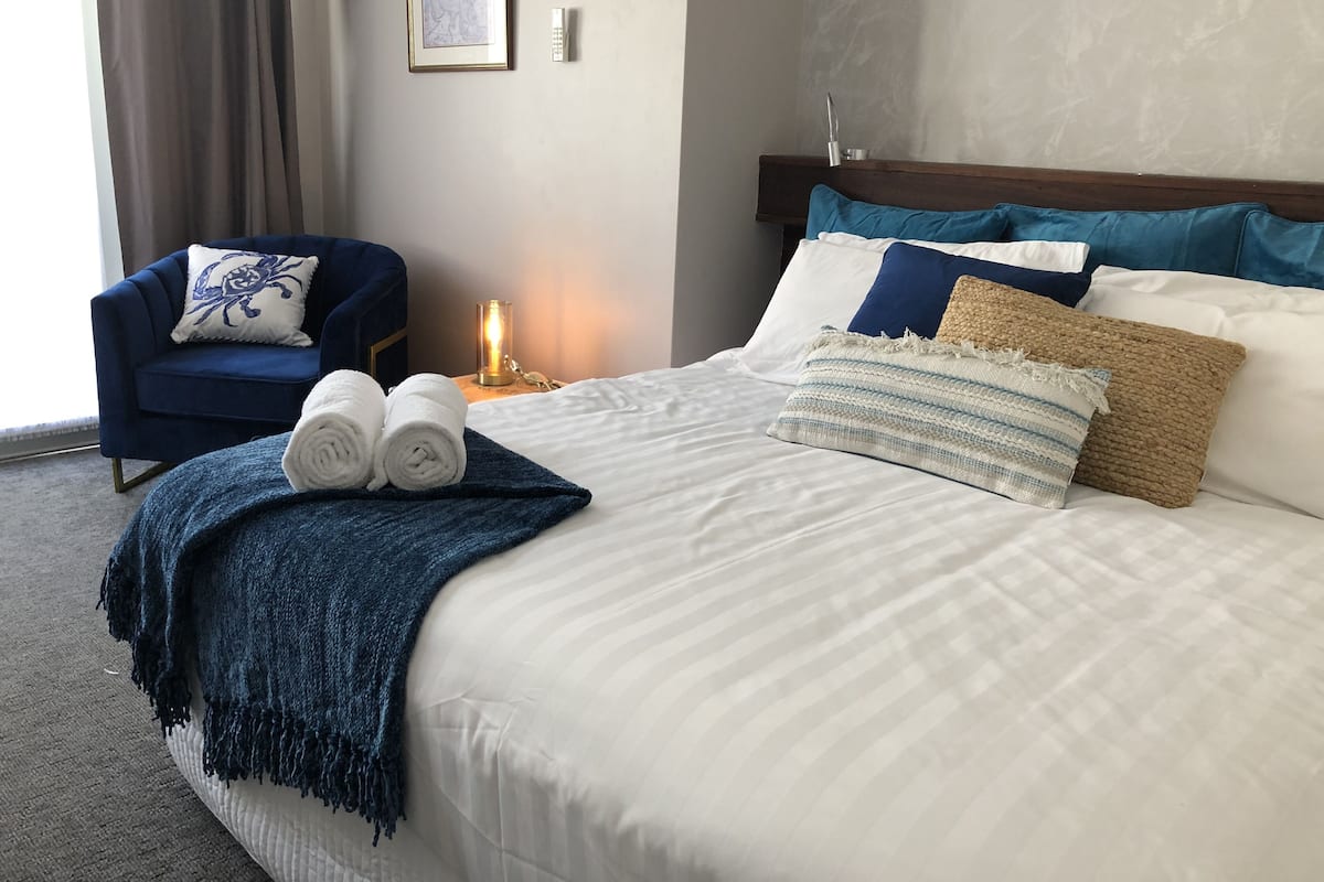 Ein ordentlich gemachtes Bett mit blauer Decke, zwei zusammengerollten Handtüchern und mehreren Kissen. Ein Nachttisch mit Lampe und einem gerahmten Bild ist zu sehen.