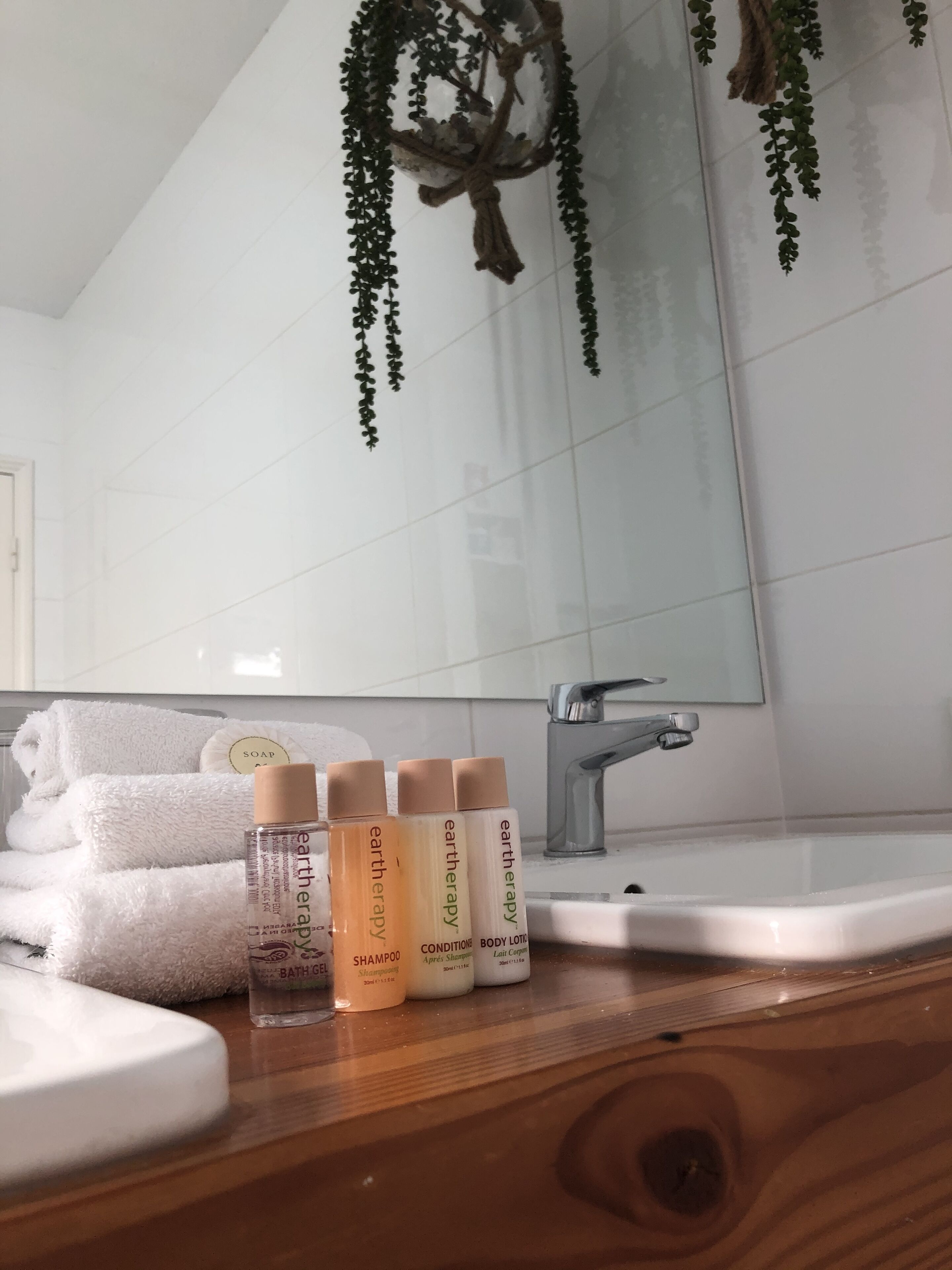 A bathroom with a wooden vanity, a white sink, and a mirror. There are three bottles of Earth Therapy body lotion and a rolled white towel on the vanity.