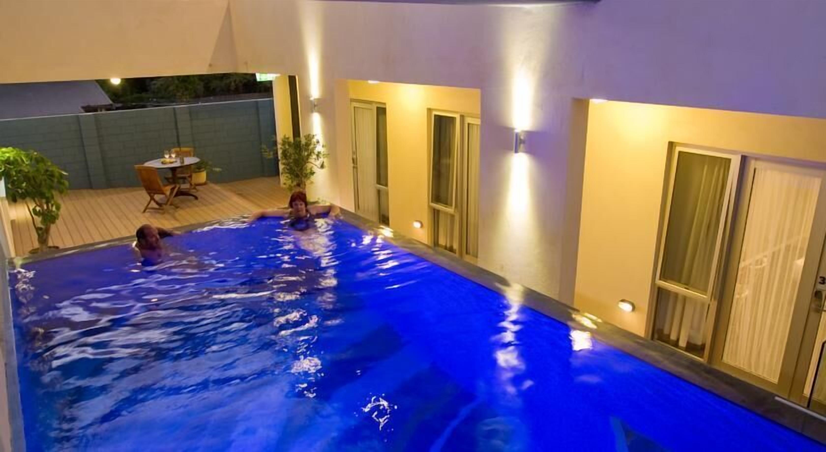 An indoor swimming pool with two people, a wooden deck, and a door with curtains.