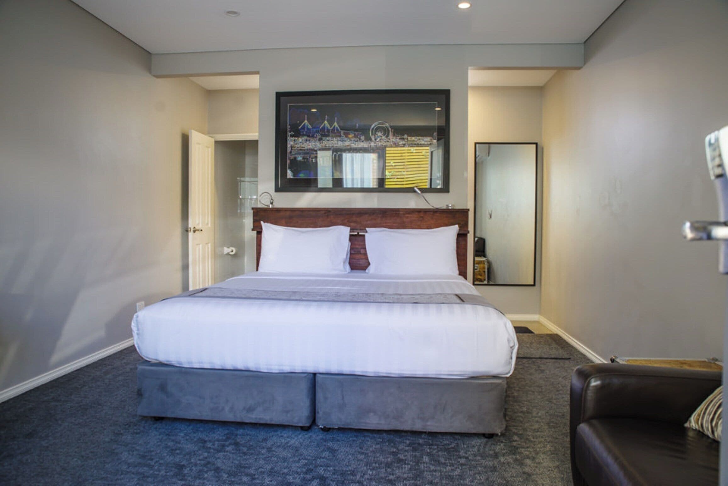 A hotel room with a large bed, a wooden headboard, a flat-screen TV mounted above the bed, and a brown leather sofa.
