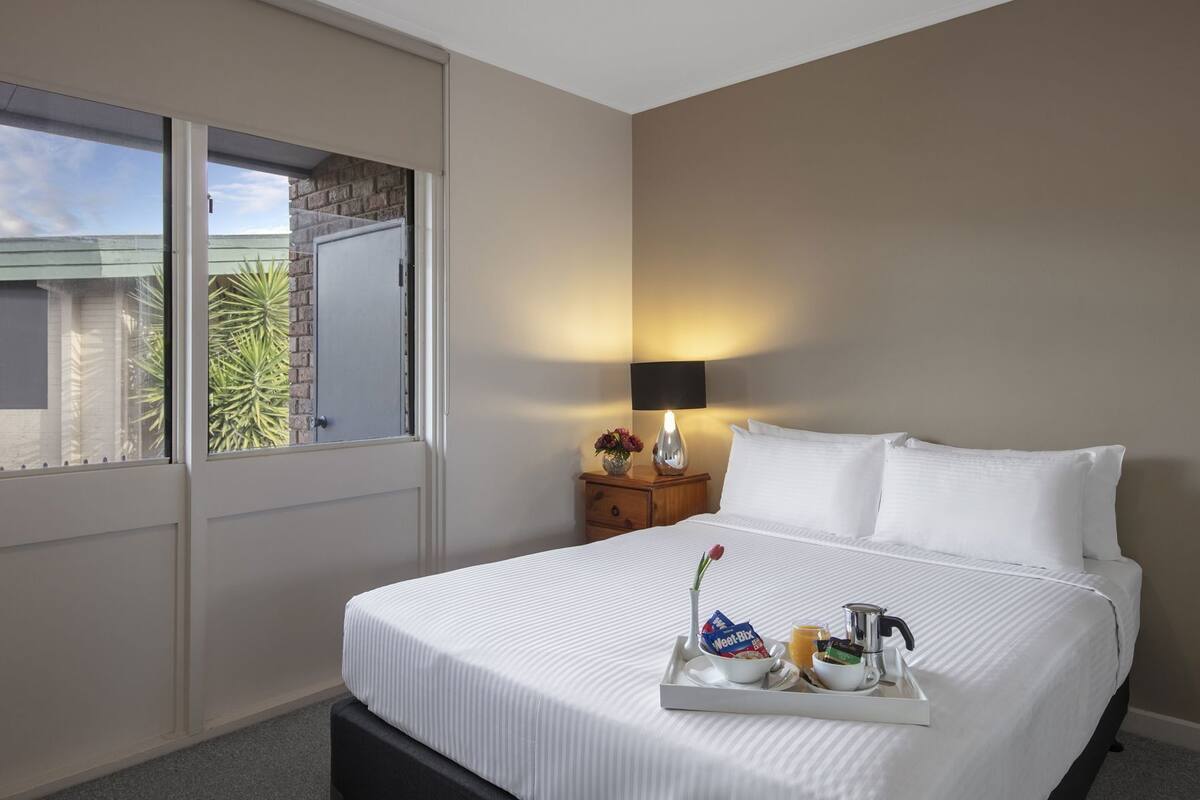 Hotelkamer met een bed, nachtkastje met lamp, dienblad met ontbijtartikelen en raam met uitzicht op groen.