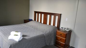 Una cama bien hecha con colcha gris, cabecera de madera, mesita de noche con teléfono y una pequeña cómoda.