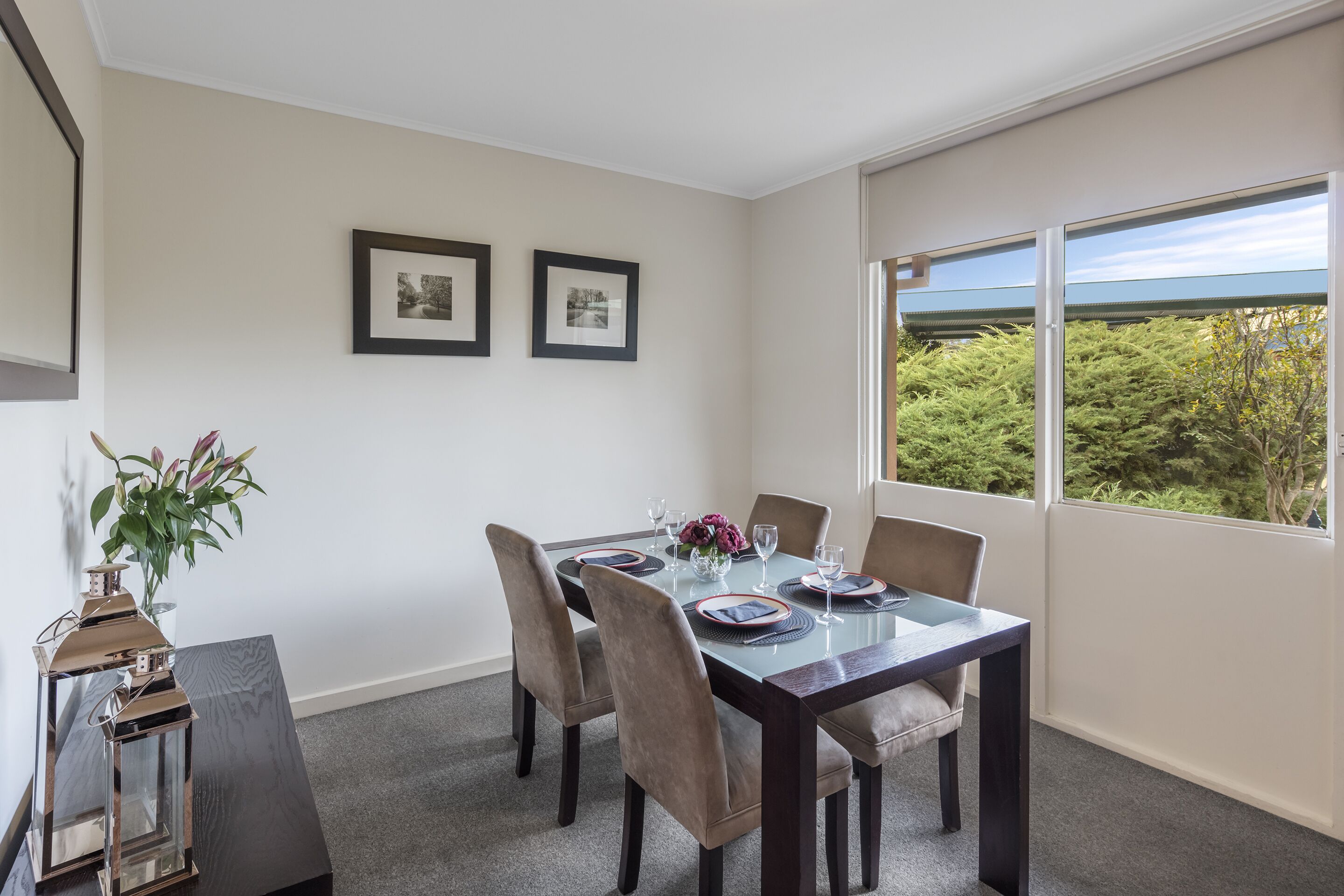 A dining area with a glass table, four chairs, and a vase with flowers. There are two framed pictures on the wall.