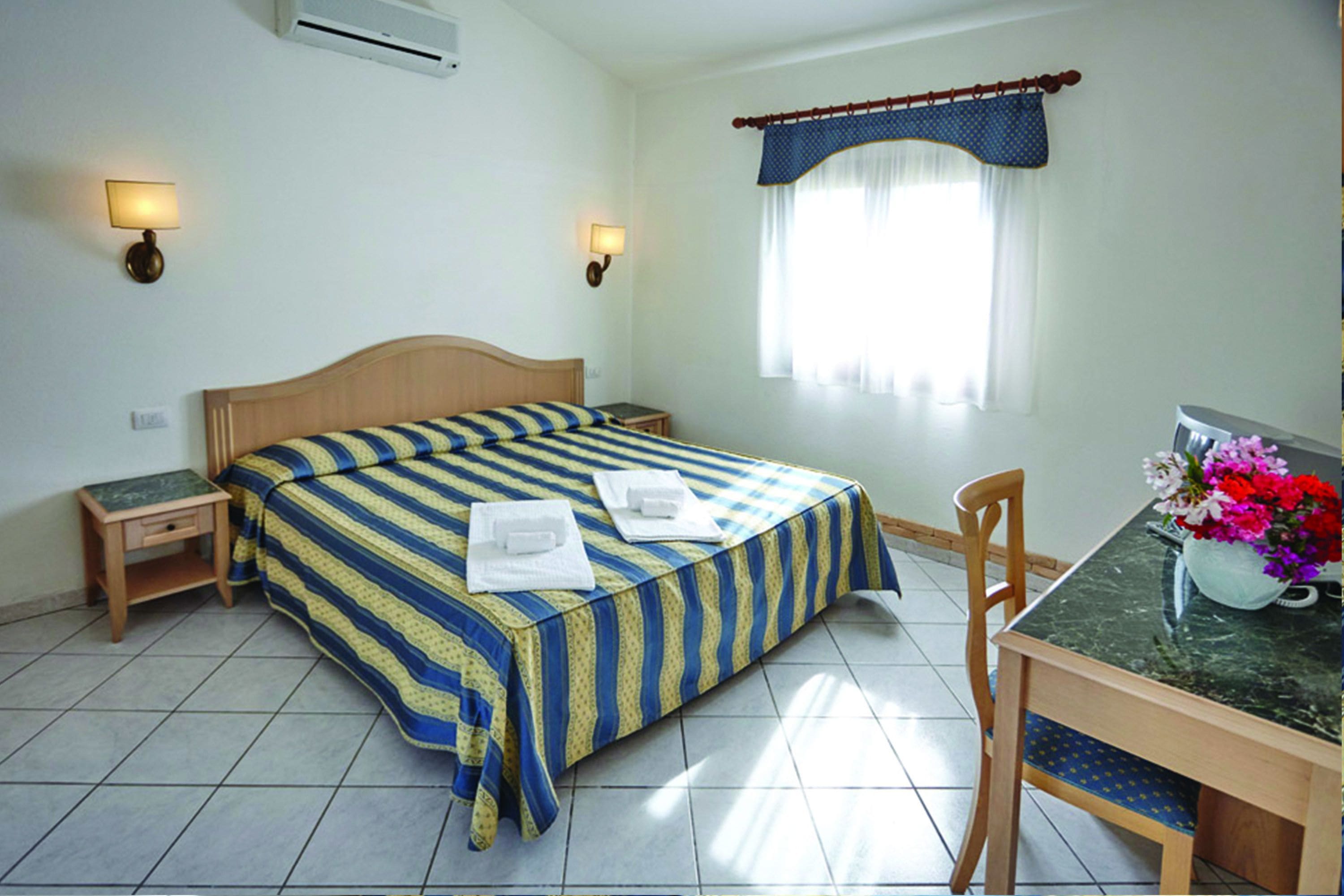 A hotel room with a bed, bedside tables, a desk with a chair, and a window with curtains.