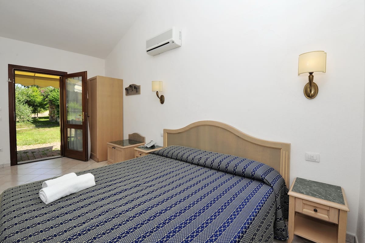 A hotel room with a bed, a nightstand, a lamp, an air conditioner, and a view of the outdoors.