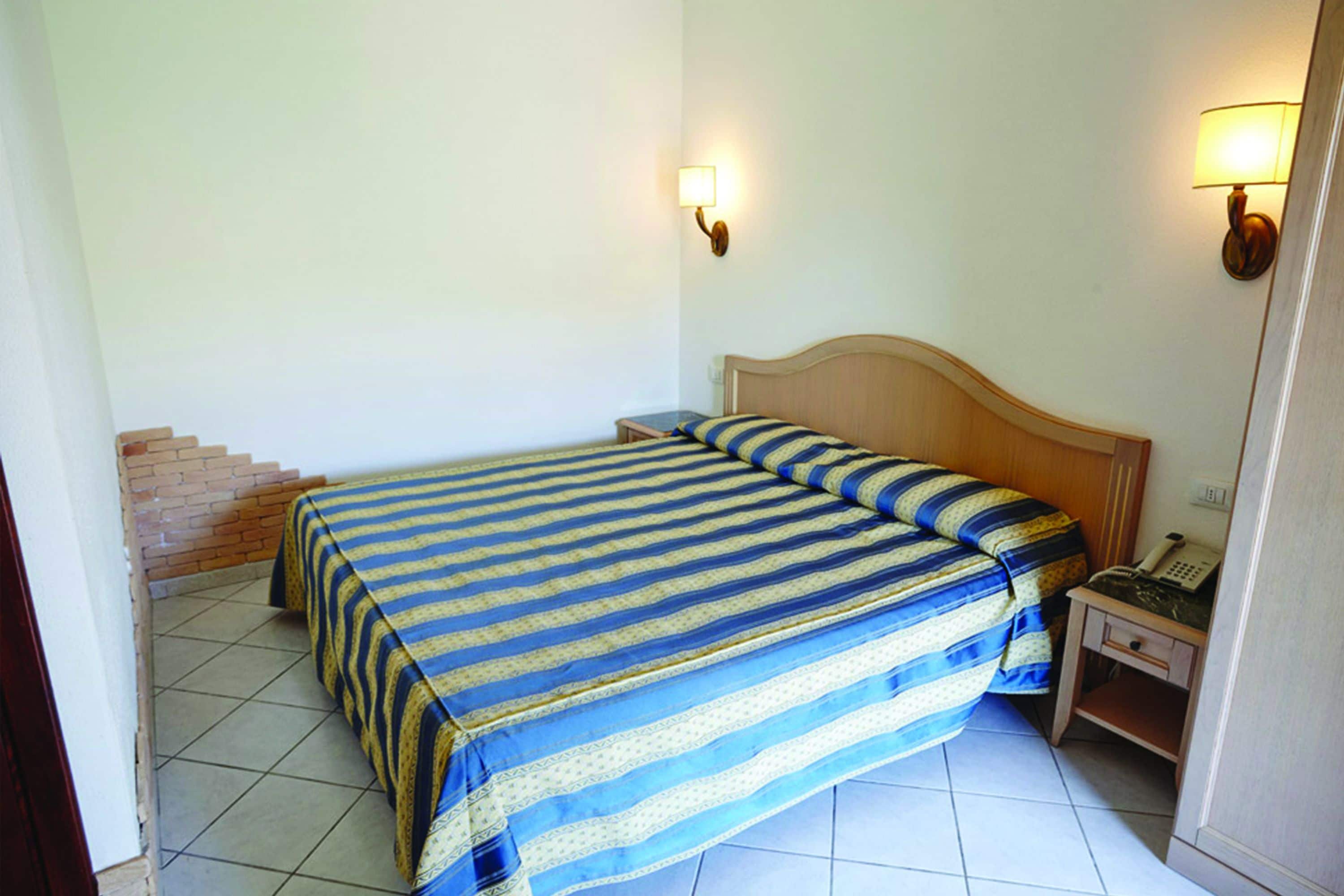 A single-bed room with a wooden headboard, striped bedding, two bedside lamps, a bedside telephone, and a small table.