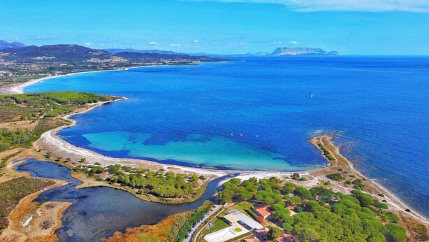 Une vue aérienne d’une zone côtière, avec une plage, des eaux d’un bleu limpide et une chaîne de montagnes à l’horizon.