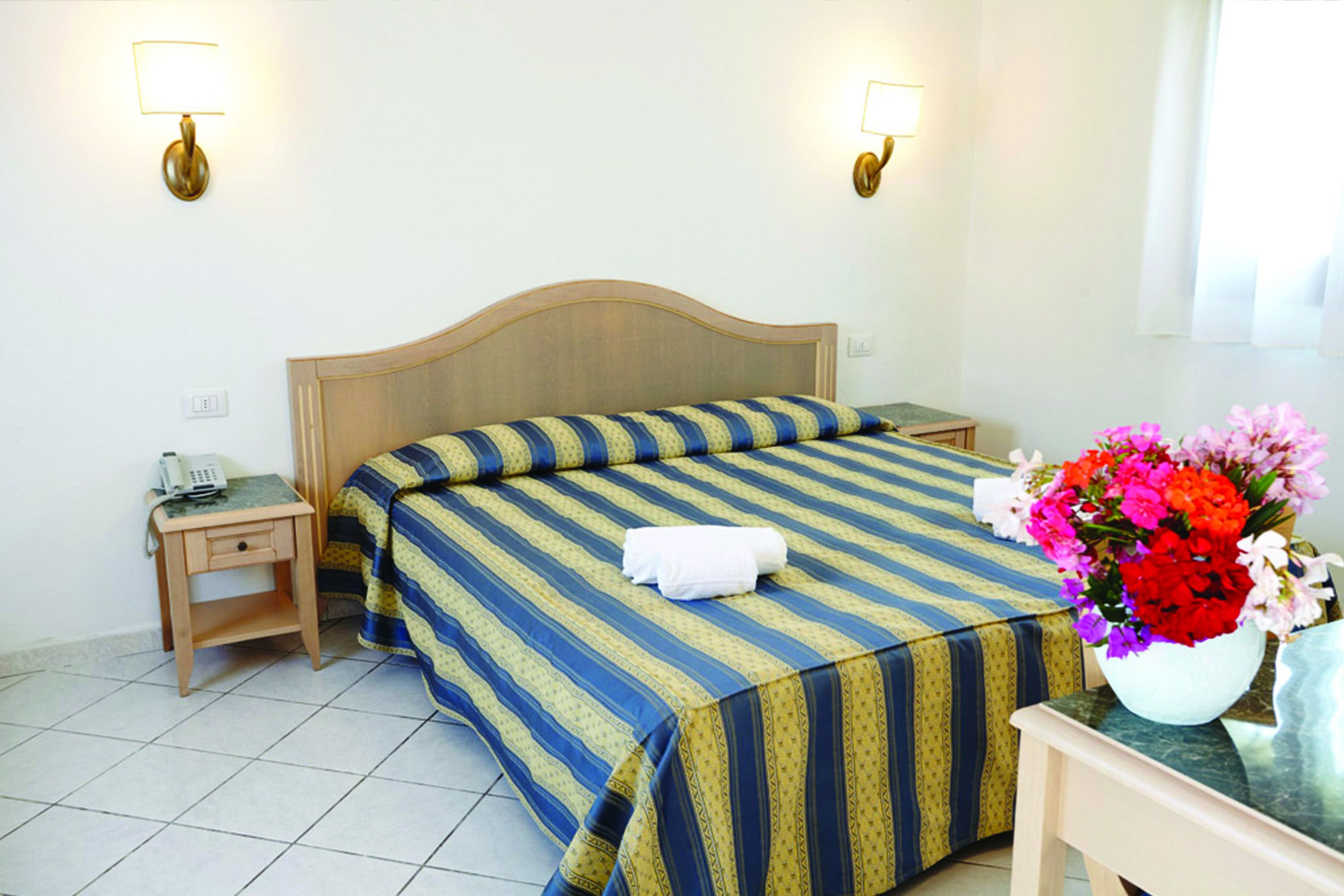 Een hotelkamer met een bed, nachtkastjes, een telefoon en een vaas met bloemen.