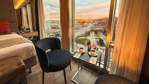 A hotel room with a bed, a chair, a small table with a cup and saucer, and a view of a cityscape through large windows. - Manesol Old City Bosphorus (Istanbul)