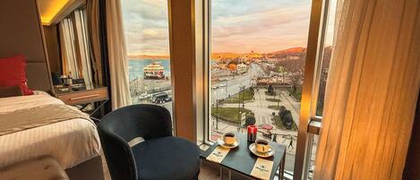 Una habitació d'hotel amb un llit, una cadira, una taula petita amb una tassa i un plat, i vista a un paisatge urbà a través de finestres grans.