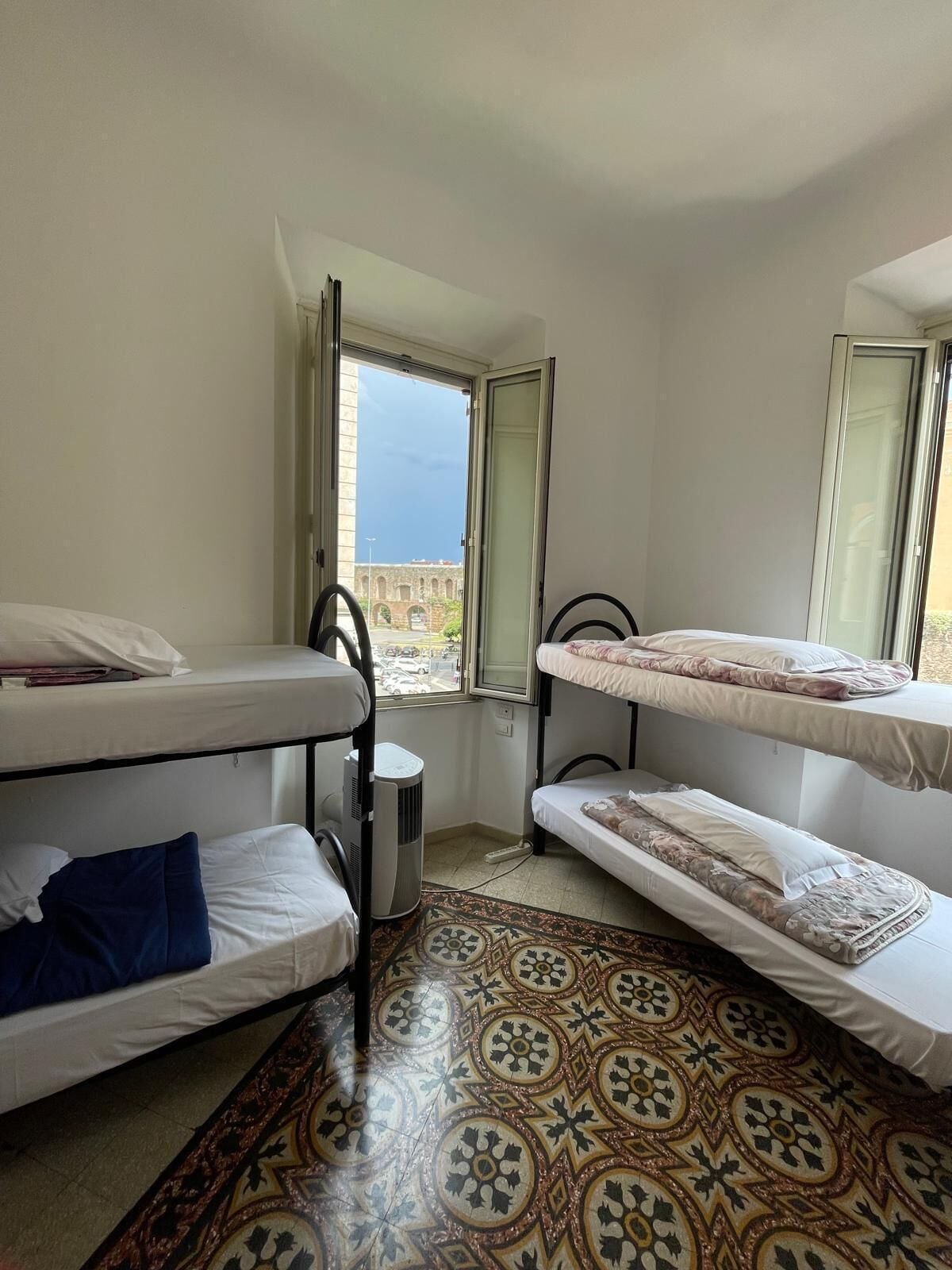 A room with bunk beds, a heater, a patterned rug, and a window with a view of a building and the sky.