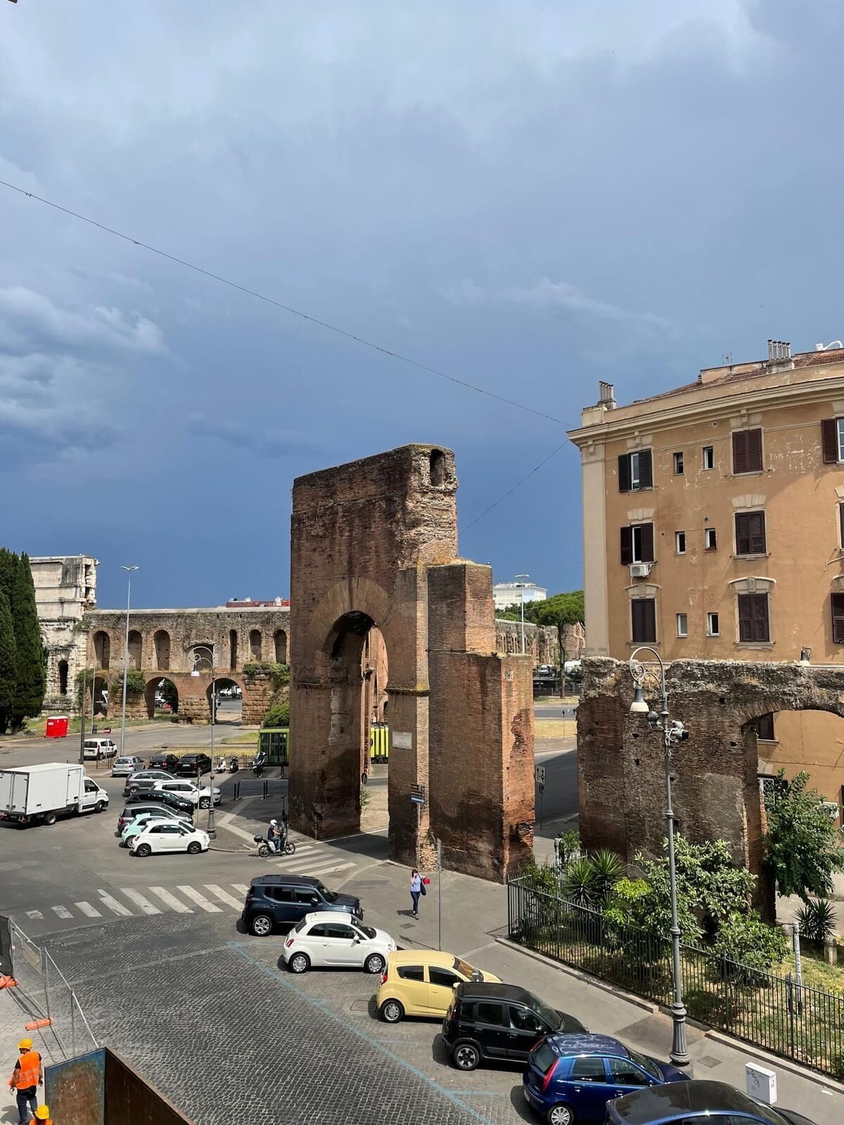 A street with a historical stone archway, modern buildings, and parked cars.