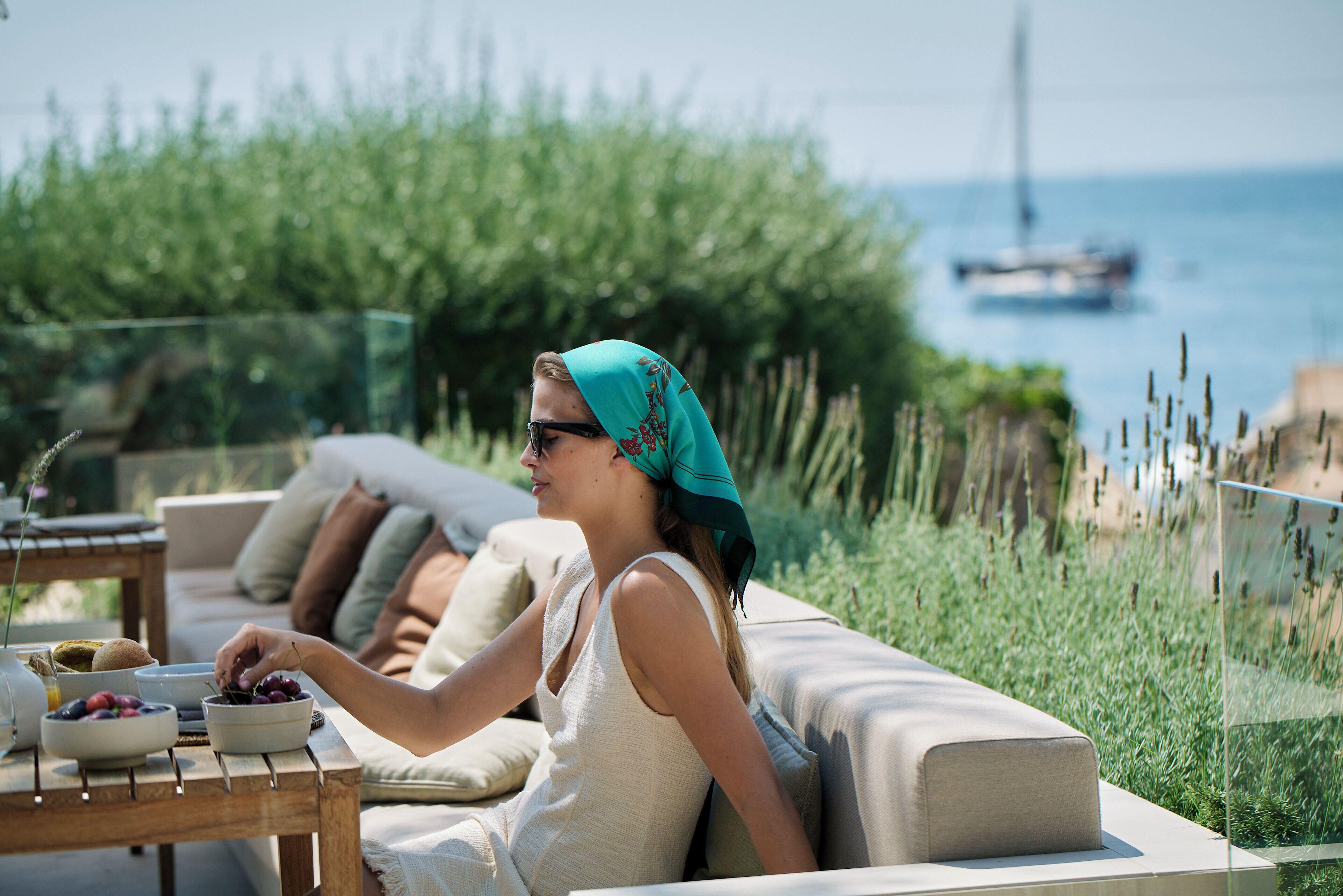 Una persona sentada en un sofá con una mesa frente a ella, un cuenco de fruta y vista al mar con barcos a lo lejos.