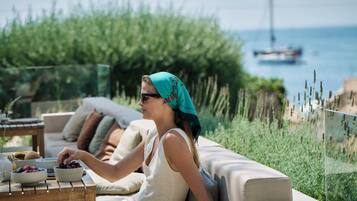 Una persona sentada en un sofá con una mesa frente a ella, un cuenco de fruta y vista al mar con barcos a lo lejos.