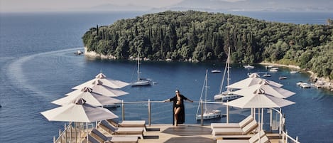 A deck with sun loungers and umbrellas overlooking a marina with boats and a forested island.