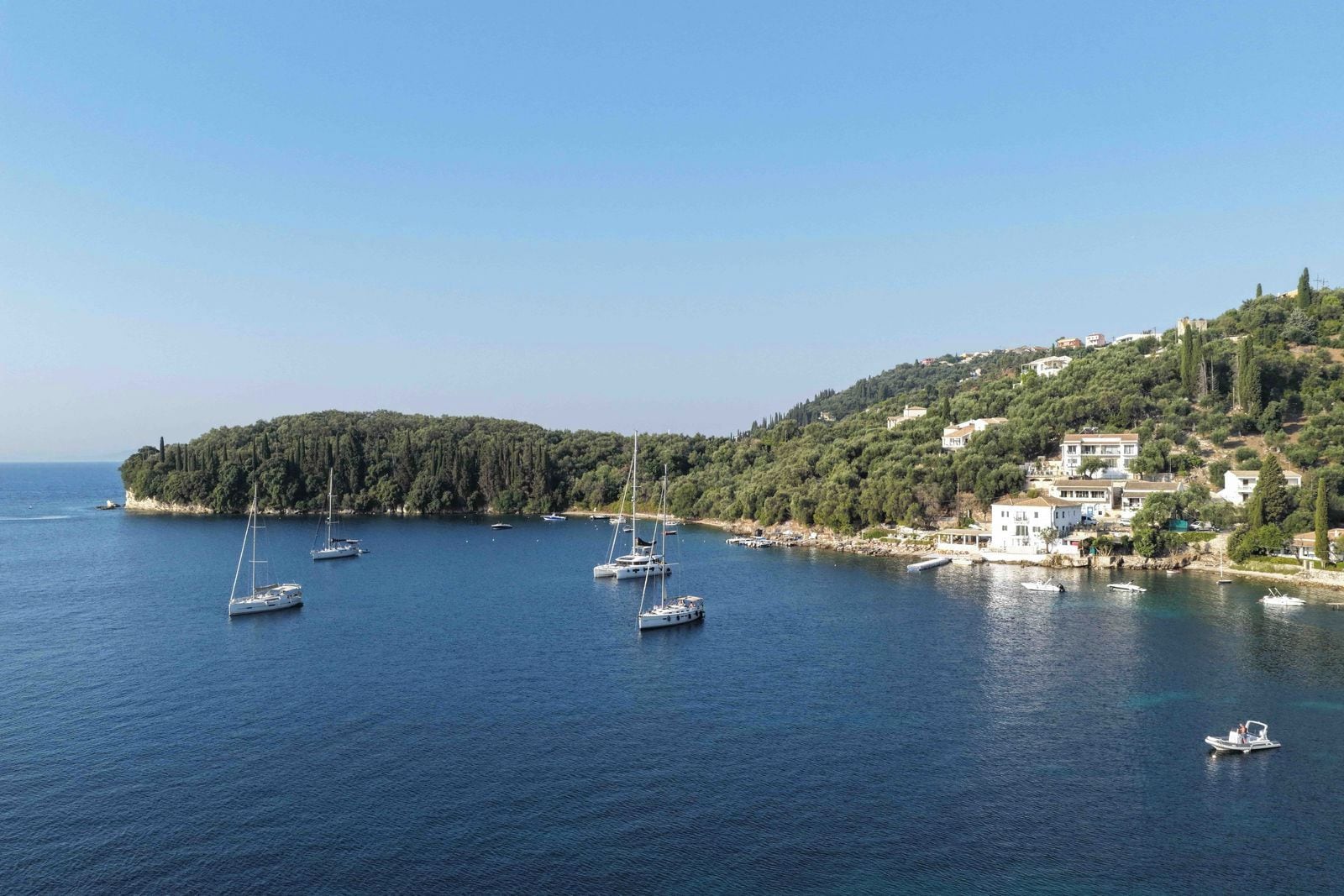 A coastal area with multiple boats anchored in the water, a hillside with buildings, and a clear sky.