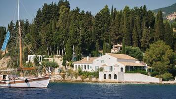 A coastal building with a white facade and a sloped roof, situated next to a body of water with a sailboat.