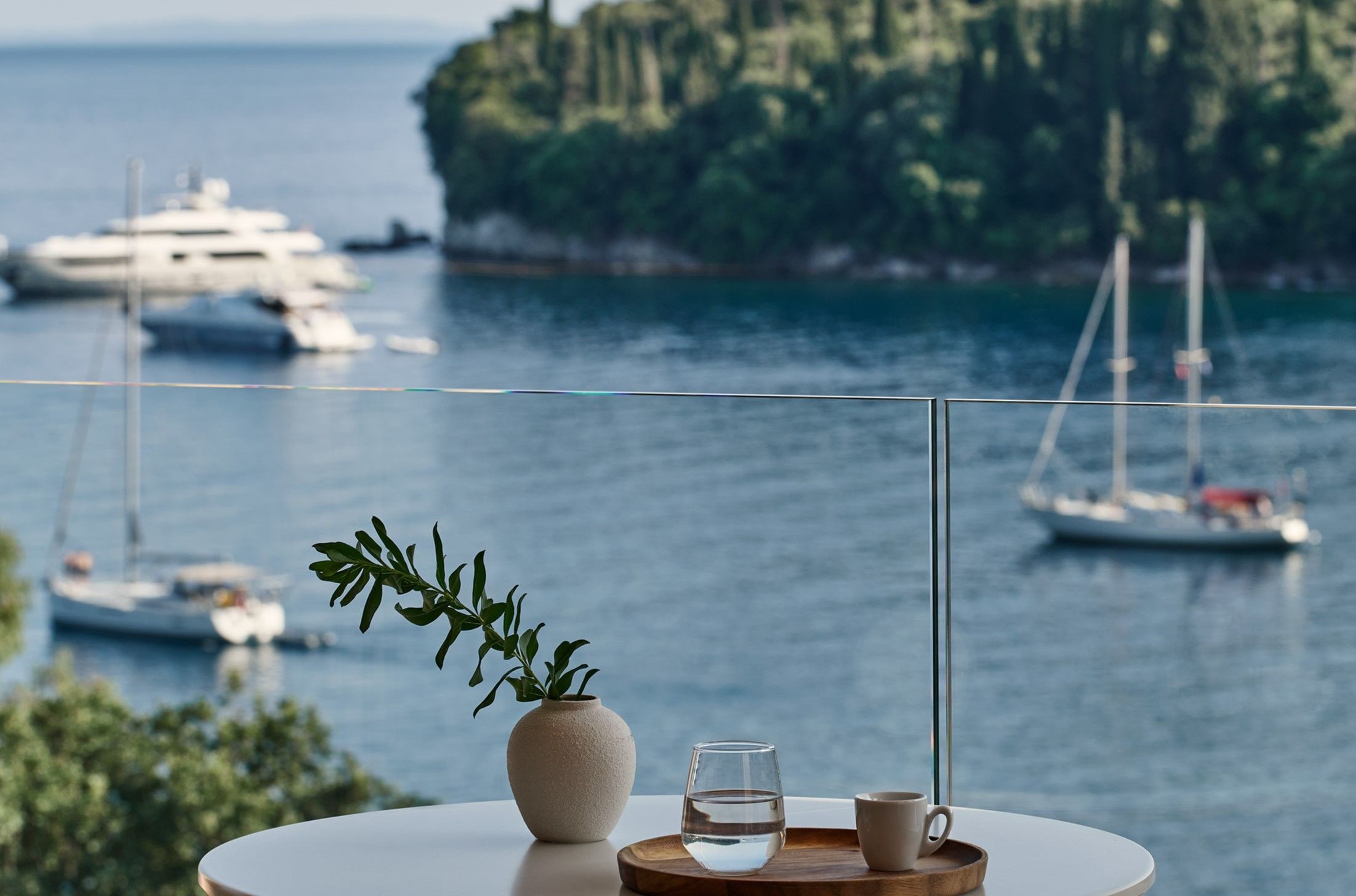 A table with a vase of green leaves, a glass of water, and a cup, overlooking a marina with boats and a forested island.