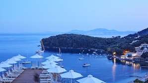 Une vue sur la côte, avec une terrasse en bois, des chaises longues et des parasols, donnant sur une marina où sont amarrés des bateaux, et sur une colline boisée.