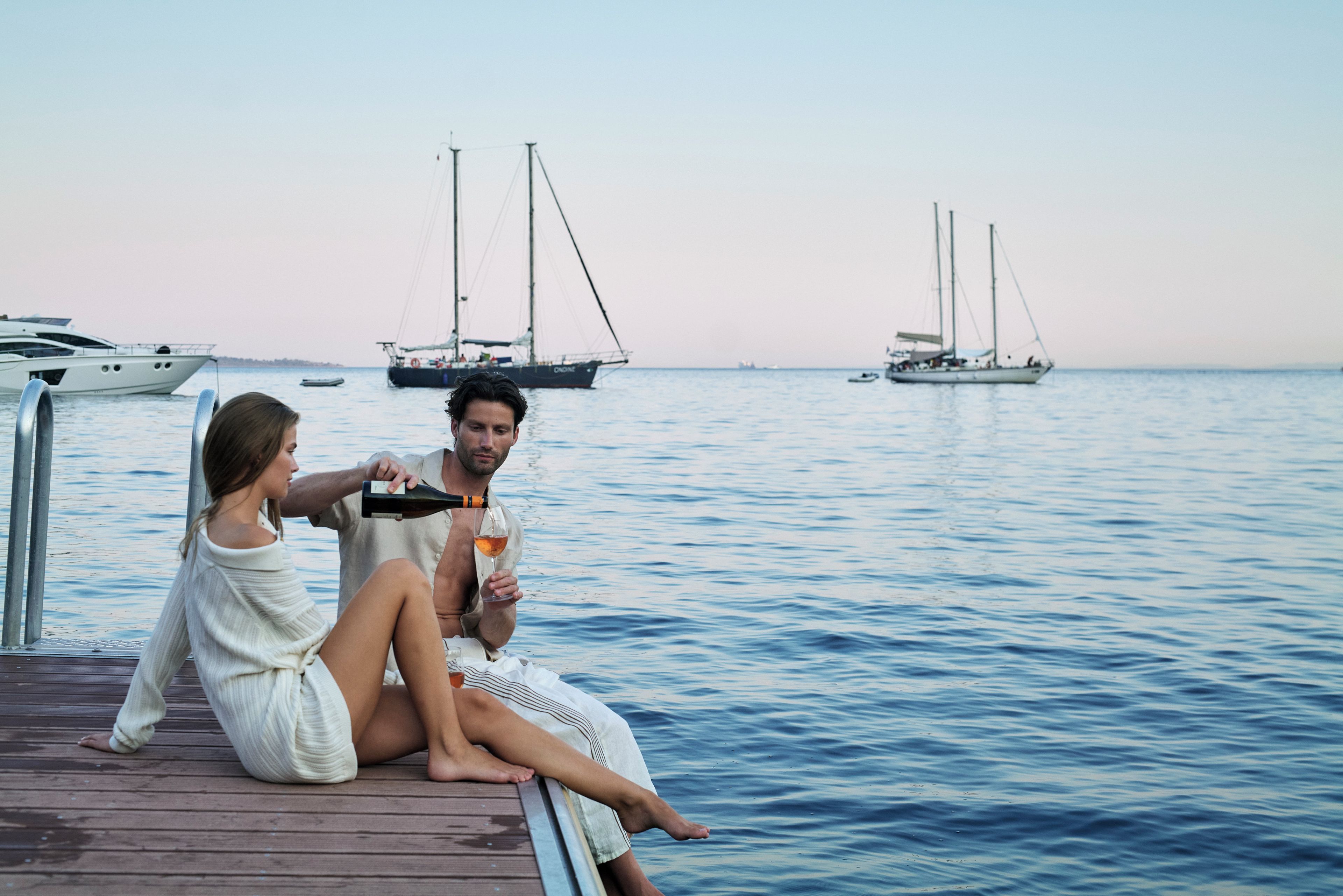 Una pareja sentada en un muelle de madera junto al mar, con veleros al fondo.