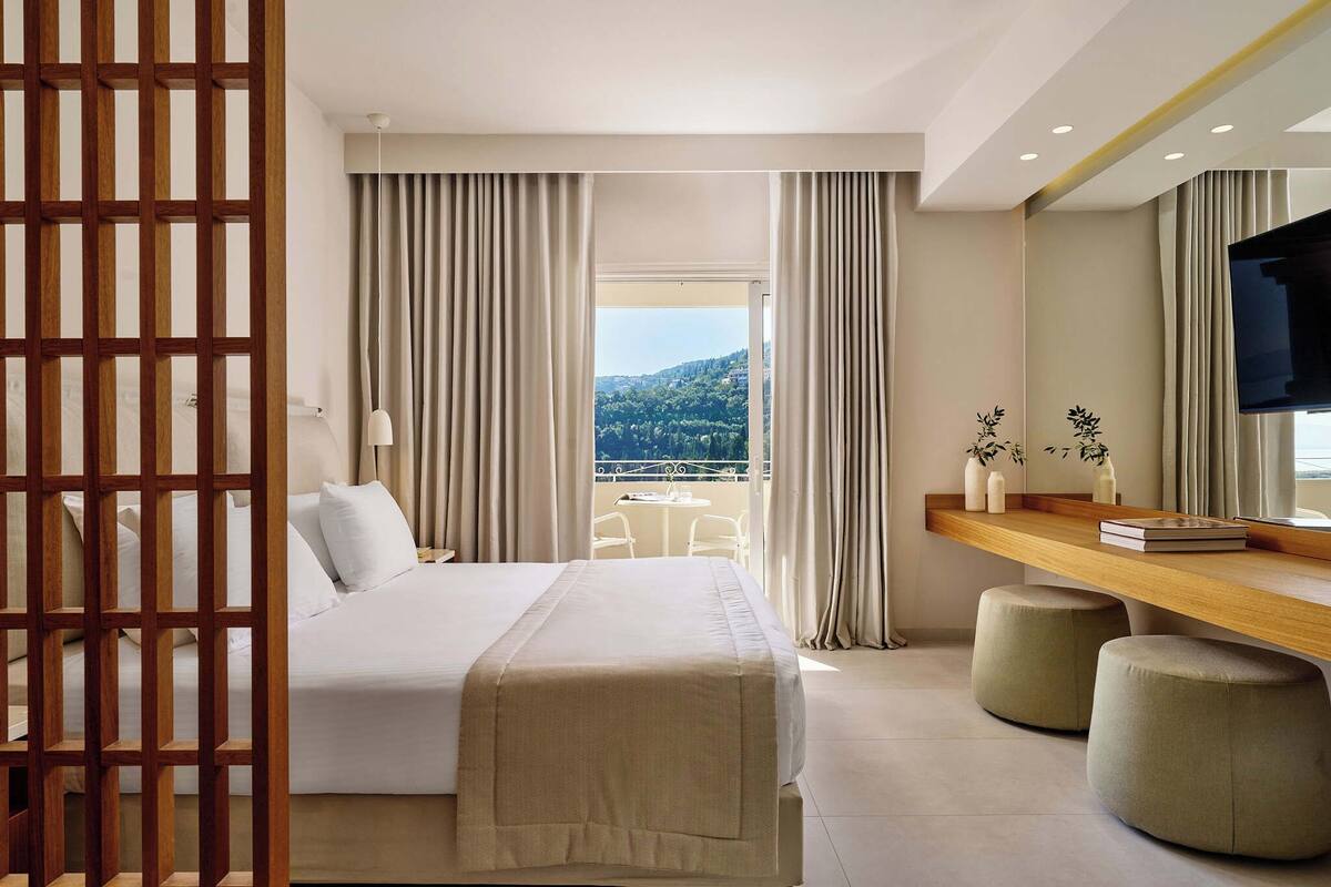 Een hotelkamer met een bed, een bureau met twee voetenbankjes, een televisie en een balkon met gordijnen.