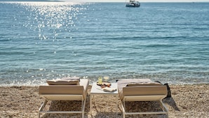 Une plage avec deux chaises longues, une table avec des boissons et des en-cas, et un voilier à l’horizon.