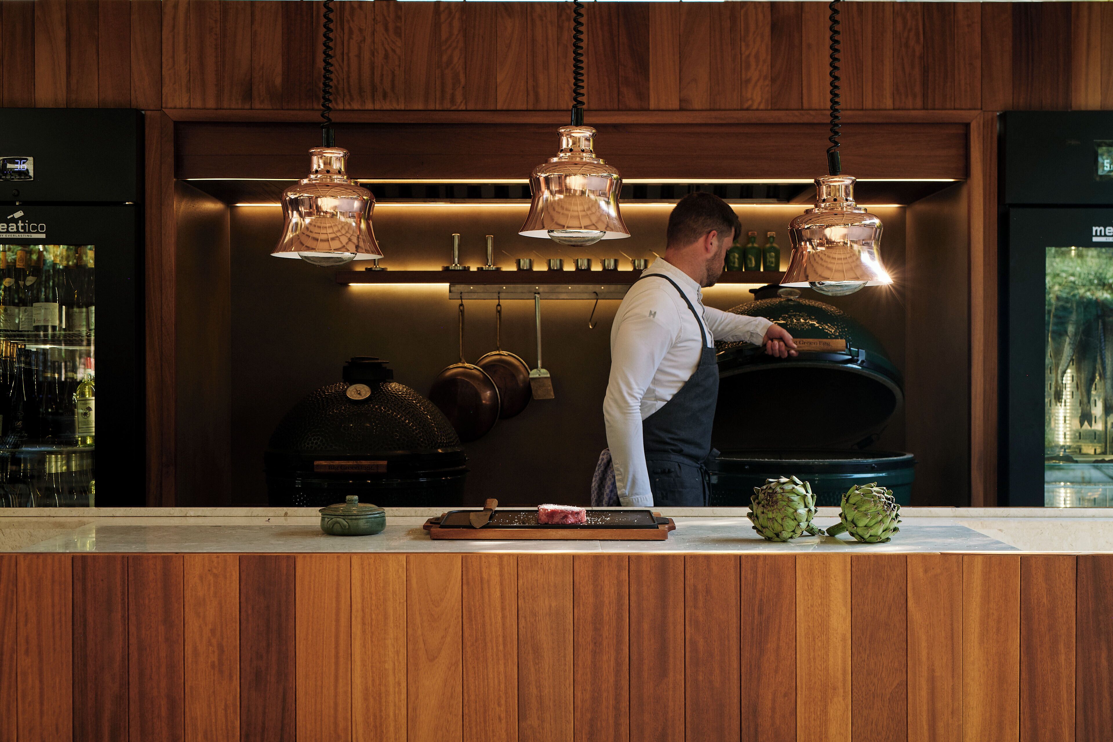 A kitchen counter with a chef preparing food, wooden cabinets, hanging pendant lights, and a refrigerator.