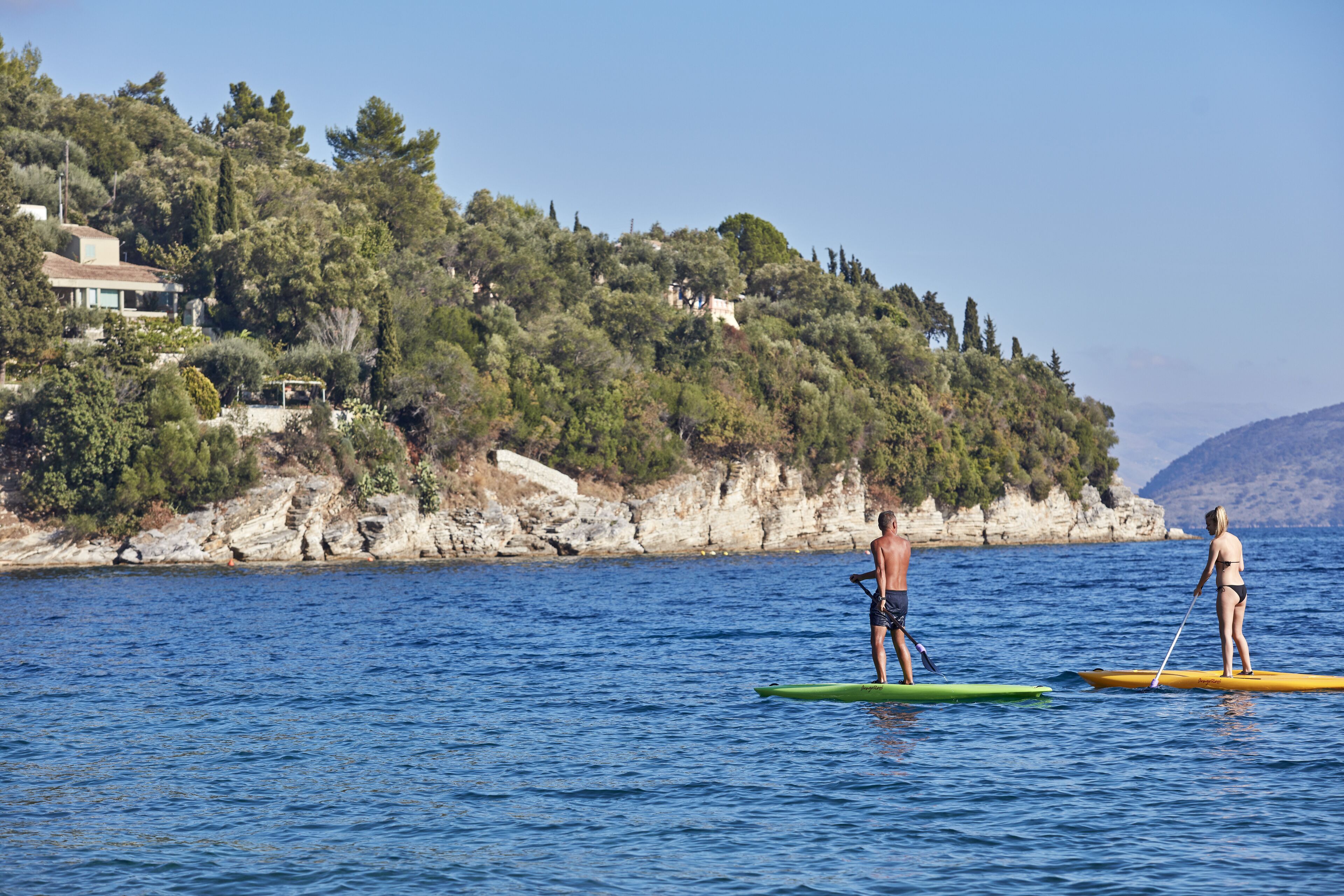 Zwei Personen stehen auf Paddleboards auf ruhiger See, im Hintergrund eine Küstenlandschaft.