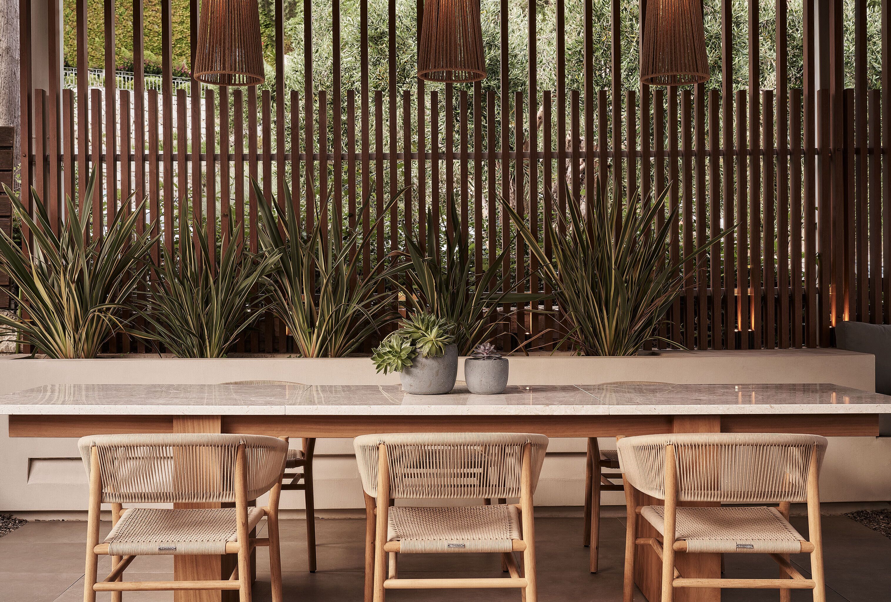 A dining area with a long table and three wicker chairs, a wooden fence, and potted plants in the background.
