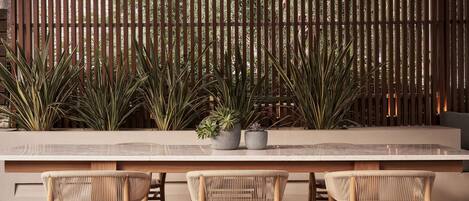 A dining area with a long table and three wicker chairs, a wooden fence, and potted plants in the background.