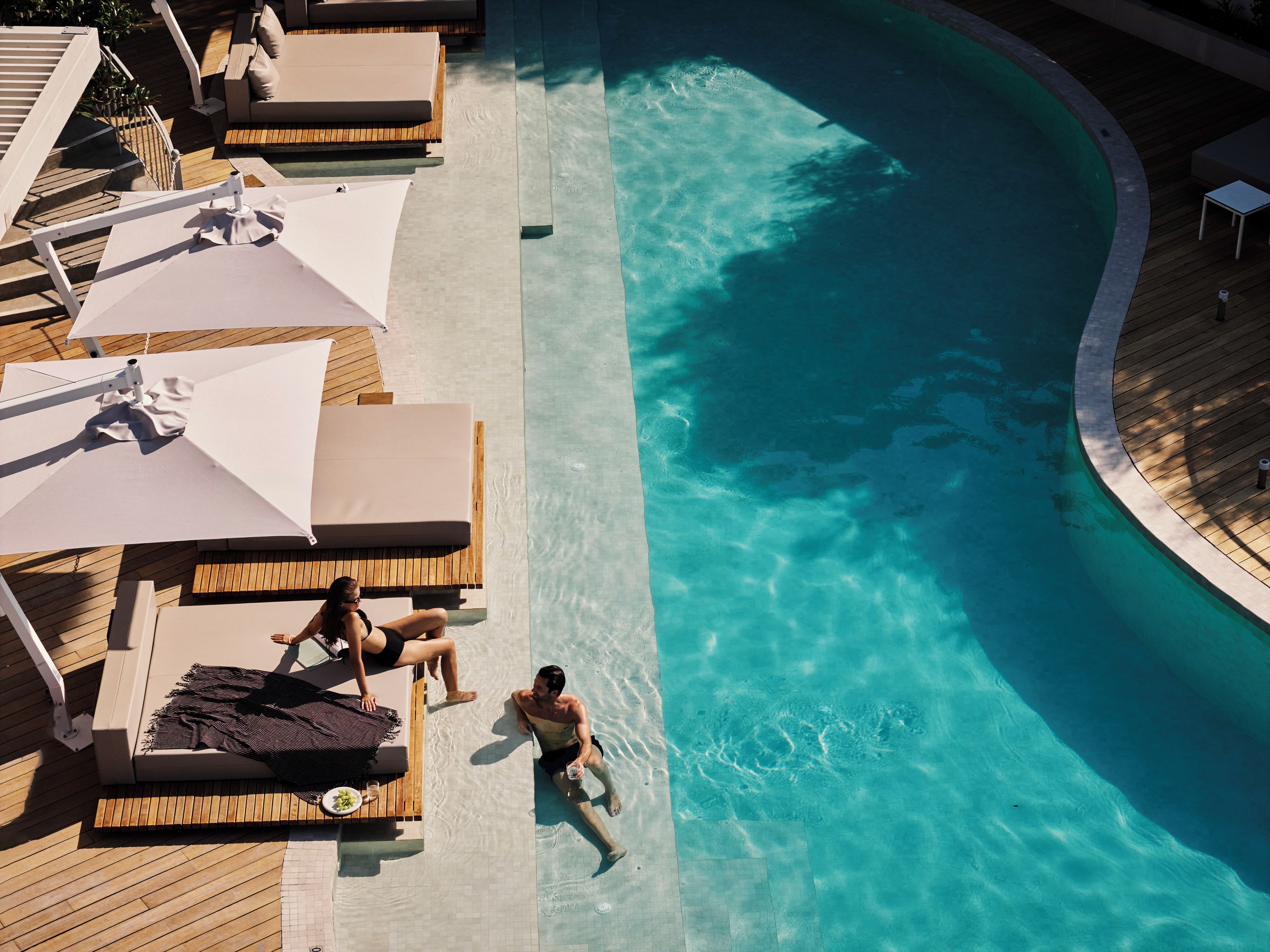 Un espace piscine aménagé avec des chaises longues et des parasols, une terrasse en bois et une piscine à l’eau d’un bleu cristallin.
