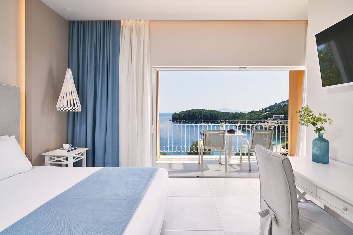 Hotelkamer met een bed, een klein tafeltje met een kopje en een boek, een stoel, en een balkon met een tafel en stoelen met uitzicht op zee.