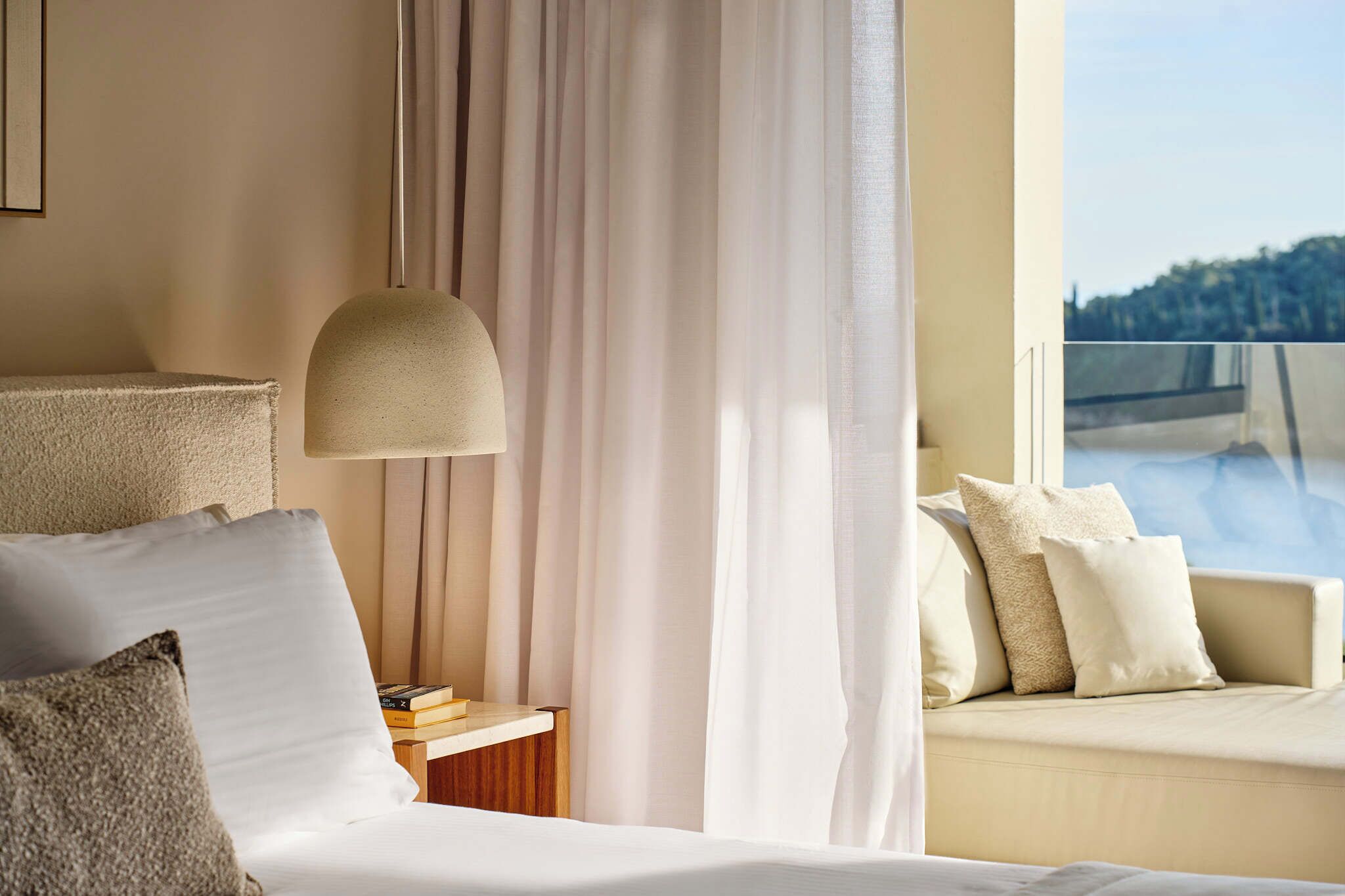 A hotel room with a bed, a bedside table with books, a lamp, a sofa with cushions, and a view of a body of water through a window.
