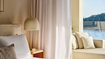 A hotel room with a bed, a bedside table with books, a lamp, a sofa with cushions, and a view of a body of water through a window.