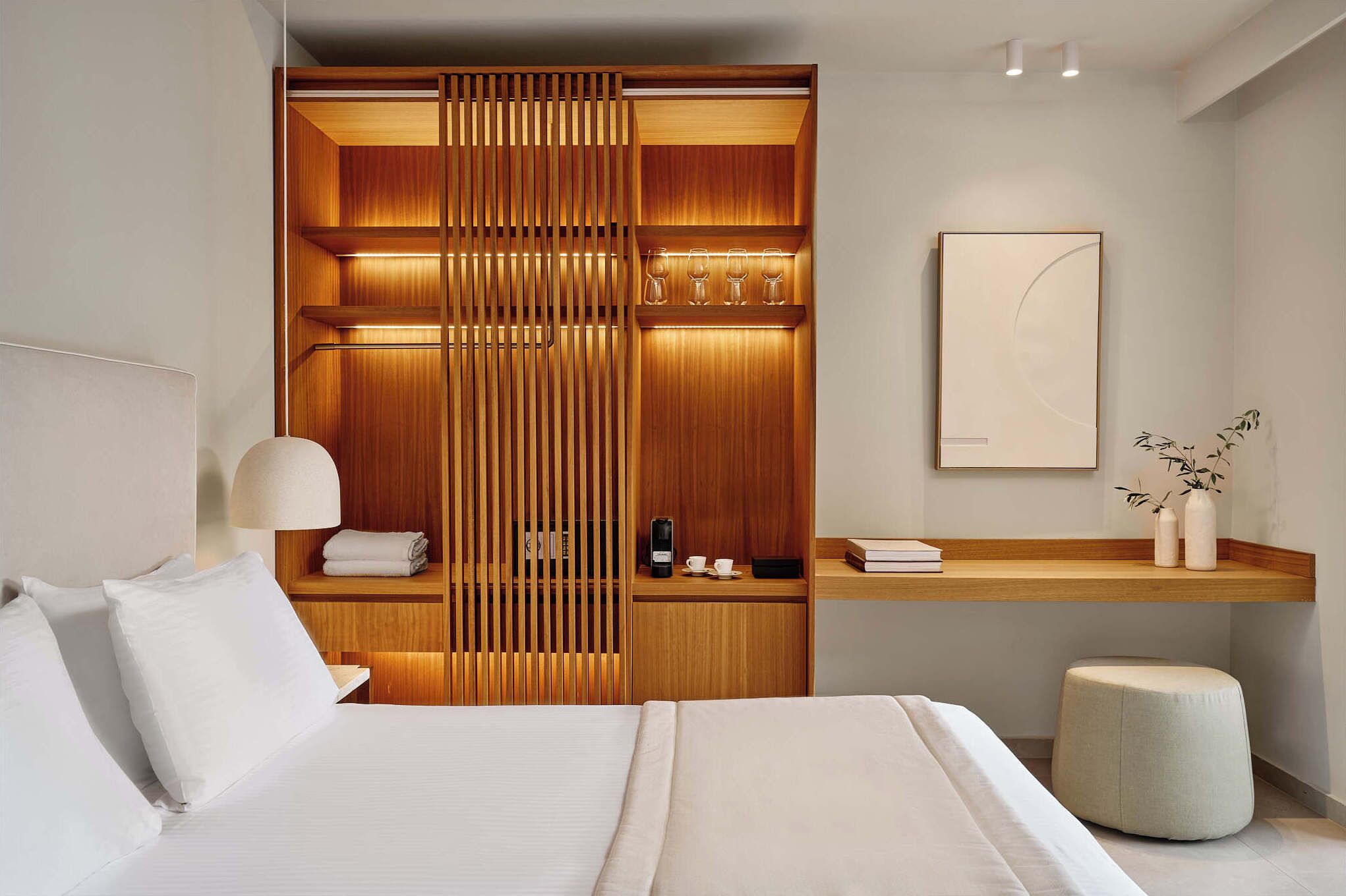 Een moderne hotelkamer met een net opgemaakt bed, een houten garderobe met ingebouwde verlichting, een nachtkastje met lamp en een klein zithoekje.