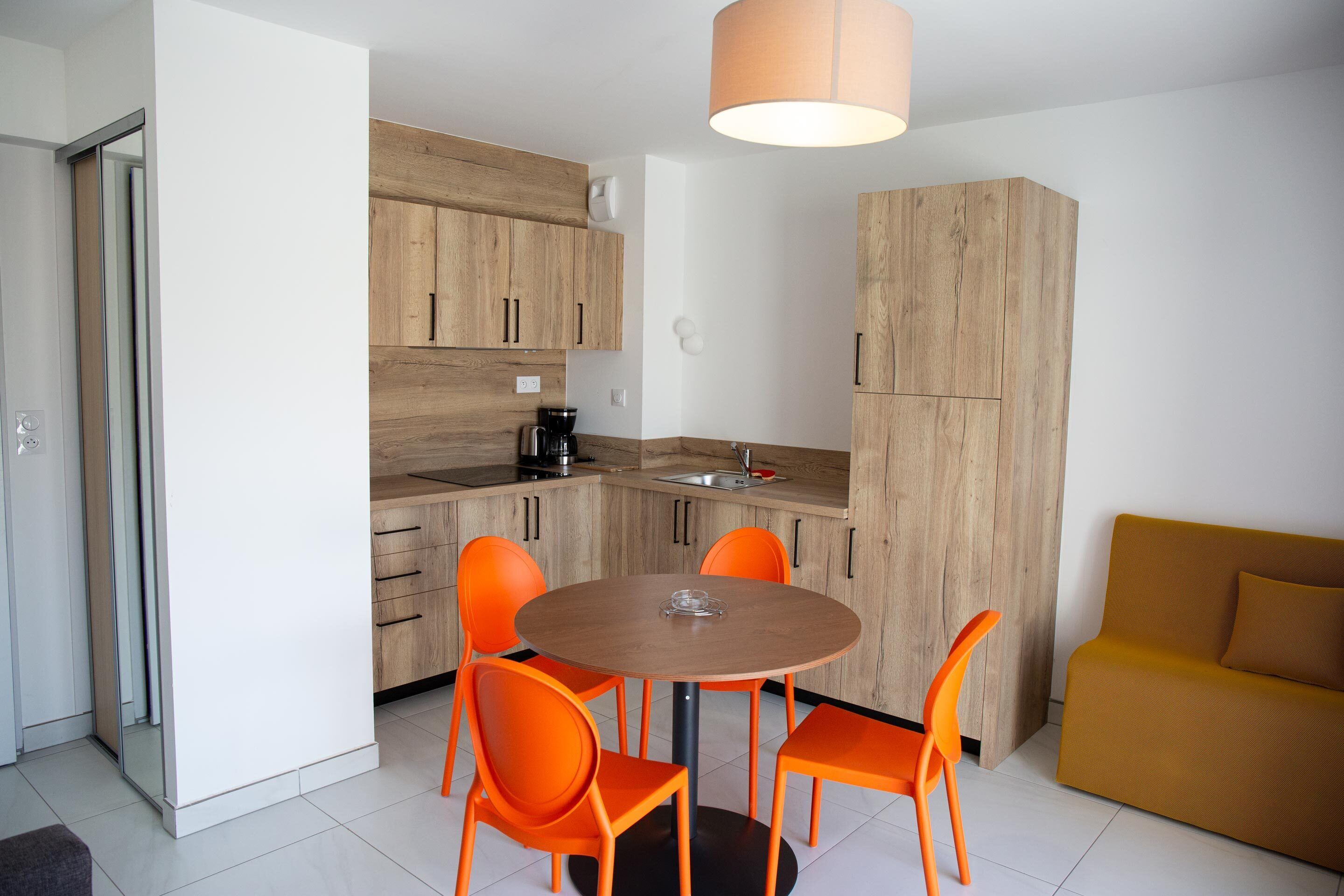 A modern kitchen with wooden cabinets, a round table with orange chairs, and a mustard-colored sofa.