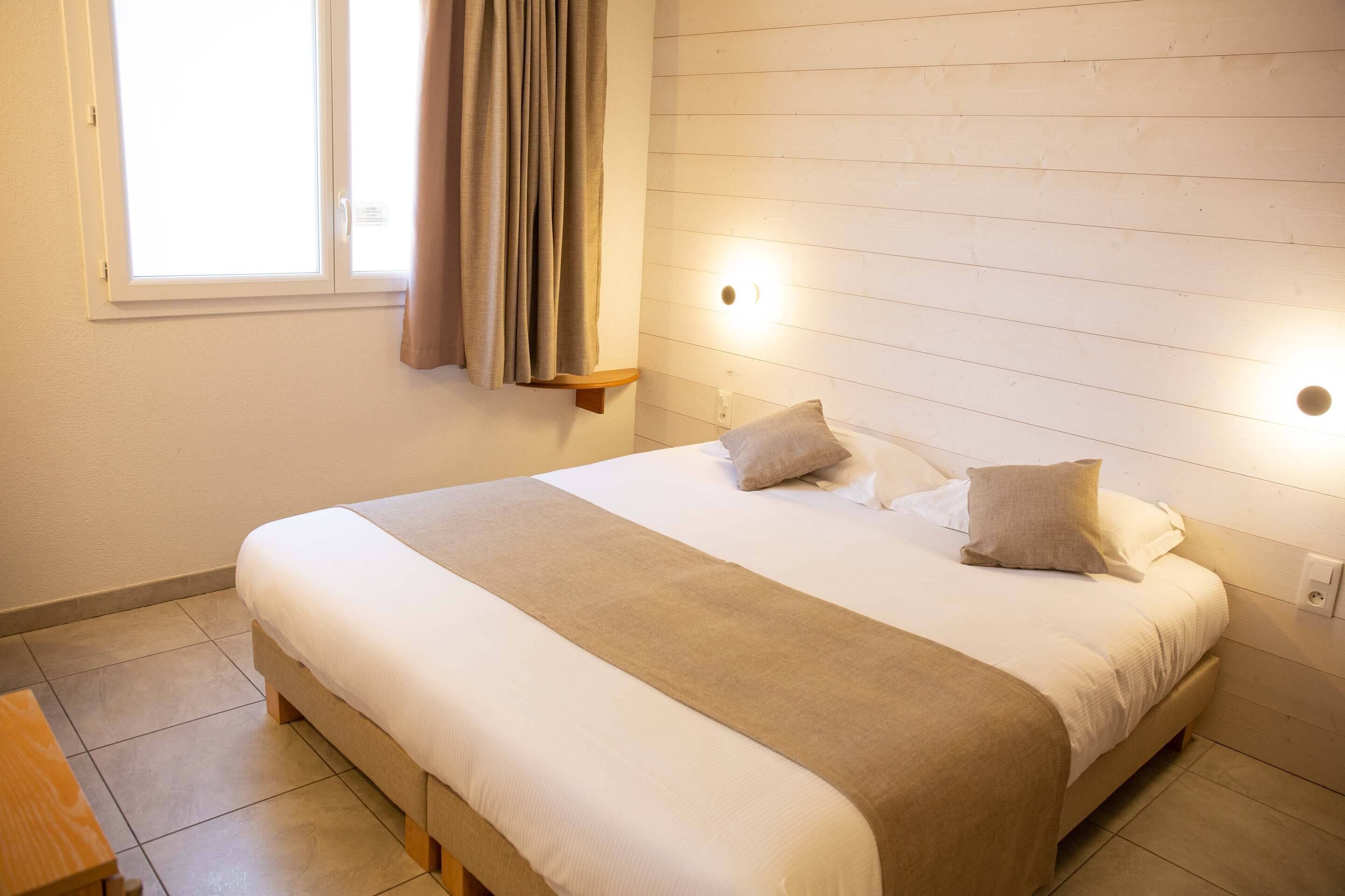 Un lit bien fait, recouvert d’une couverture beige, une table de chevet en bois et une fenêtre avec des rideaux.