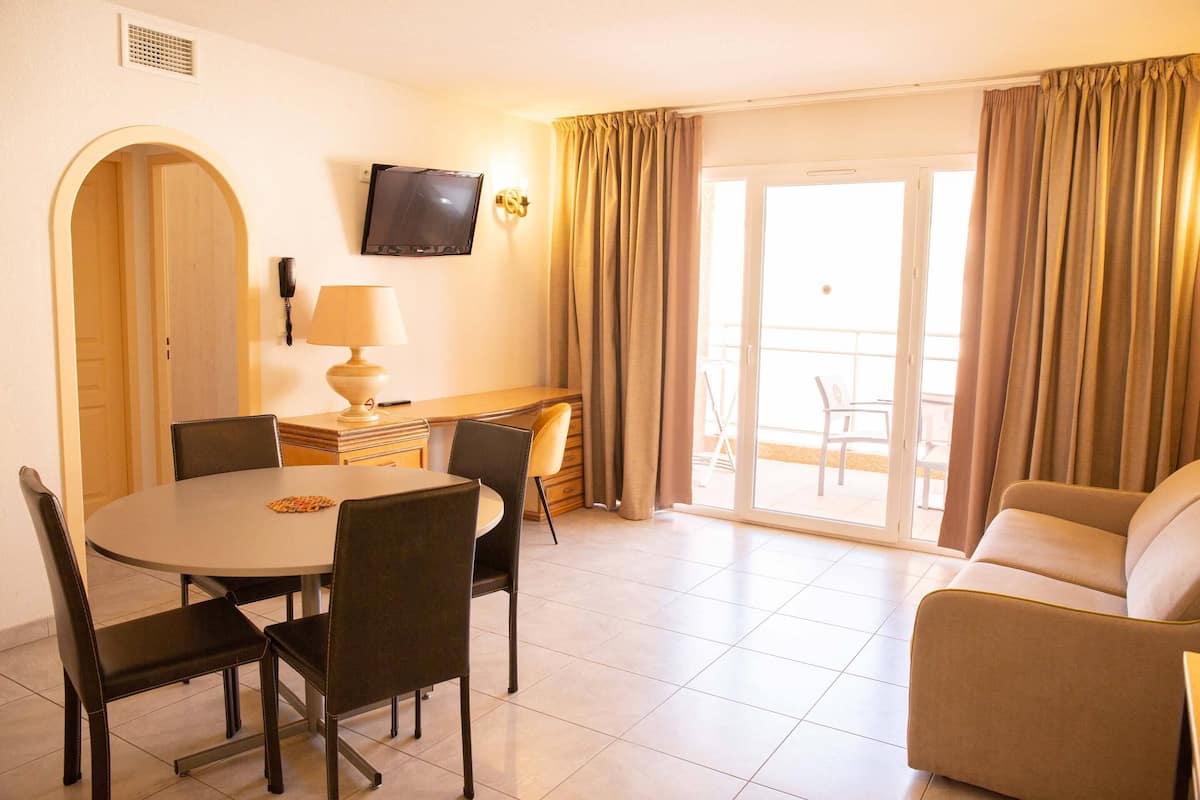 A hotel room with a dining area, a sofa, a TV, and a balcony with curtains.