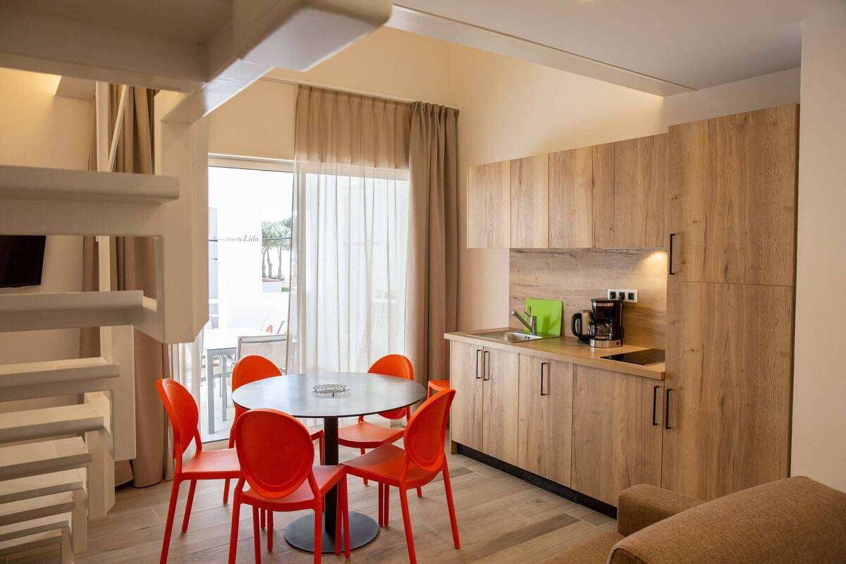 A modern kitchen with wooden cabinets, a round table with orange chairs, and a coffee maker.